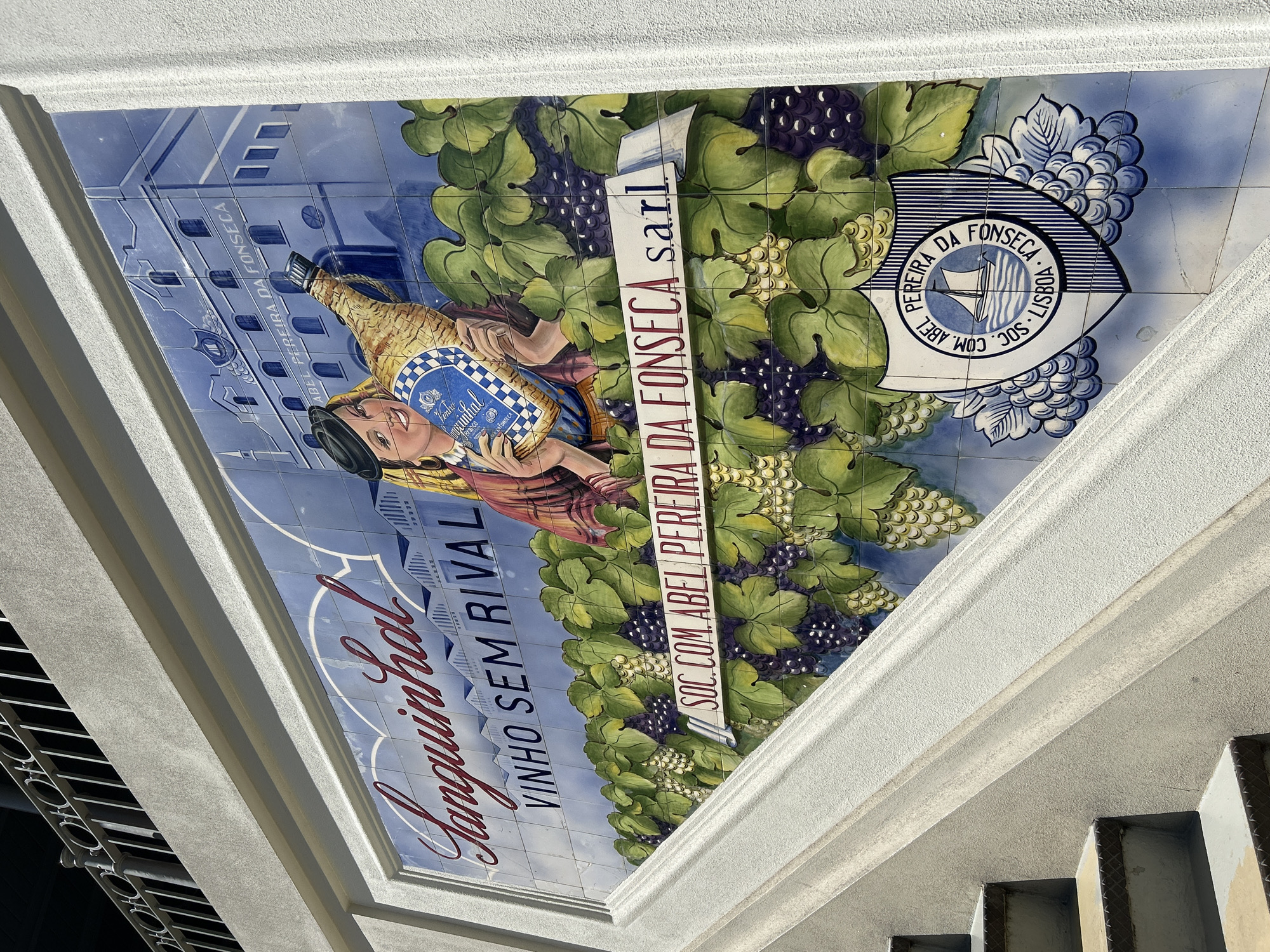 This is a photograph of a tile mural decorating an exterior wall, possibly of a building or a historical site. The mural depicts a bucolic scene with a strong emphasis on grapes and wine.
The central subject is a woman holding a bottle of wine, identifiable by its shape and the label. She appears to be dressed in a red and pink garment, with a black hat and a straw basket. Surrounding her are abundant clusters of purple and light green grapes, interwoven with large green leaves.
To the left of the woman, a building with several windows is depicted, rendered in blue and white tiles. Above the building, text is visible on a light-colored banner: "PEREIRA DA FONSECA".
To the right, a crest or emblem is featured, also rendered on tiles. It's circular with a nautical theme in the center, a stylized sailboat on water, enclosed by a shield-like design. Text around the crest reads: "PEREIRA DA FONSECA", "SOC. COM. ABEL", and "LISBOA".
Below the main scene, large, stylized lettering in red reads "Sanguinhal VINHO SEM RIVAL", translating to "Sanguinhal Wine Without Rival".
The overall impression is that of an advertisement or dedication to a wine brand, "Sanguinhal", and possibly the "Pereira da Fonseca" company, possibly from Lisbon. The mural is bright and colorful, suggesting it's a sunny day. The composition is vertical, suggesting it might be part of a larger facade or a decorative element on a building. The style of the tiles and the artwork suggests a traditional or historical context, likely in Portugal given the mention of Lisbon.