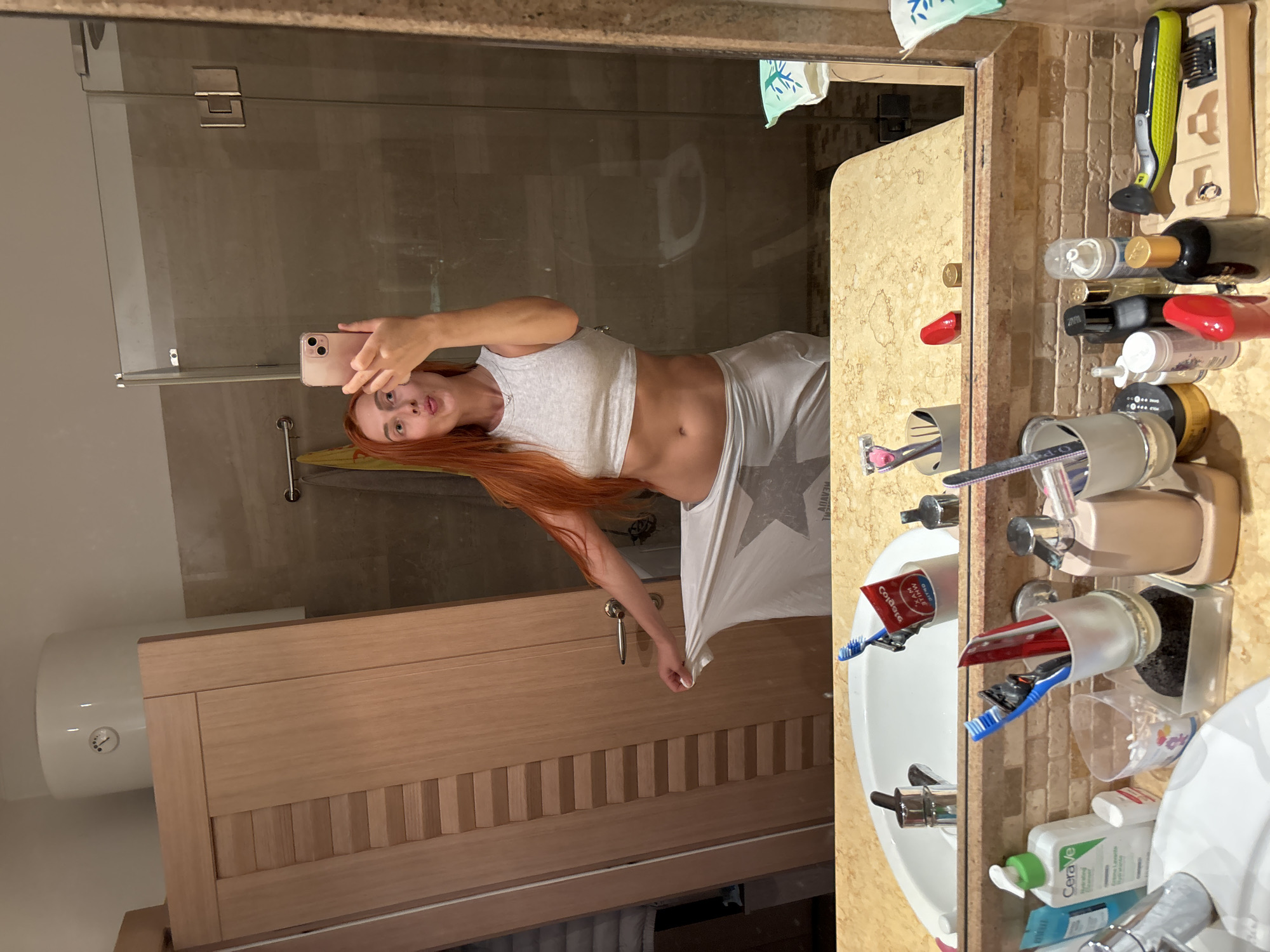 In a modern bathroom with beige tiled walls and a granite countertop, a woman with long red hair is taking a mirror selfie. She is wearing a grey crop top and a white skirt. She is puckering her lips and holding a pink smartphone in her right hand. To her left, a wooden door with horizontal slats is visible, and a white cylindrical water heater is mounted on the wall above it. The countertop is cluttered with various toiletries, including toothbrushes, toothpaste, razors, and bottles of lotion and perfume. A white sink with a chrome faucet is in the foreground, and a large mirror reflects the scene. The bathroom appears to be in an apartment or hotel room in Budva, Montenegro.