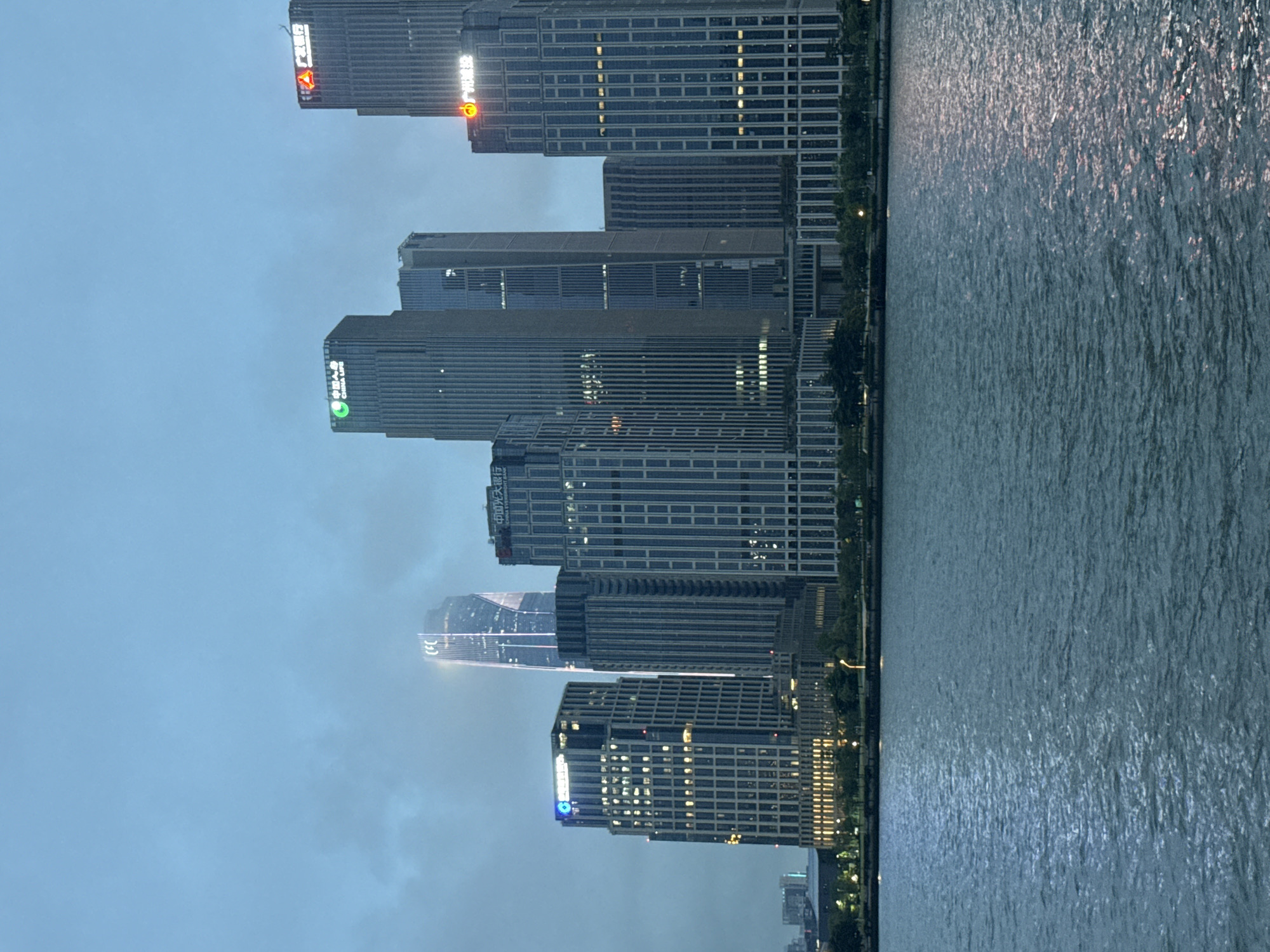 This media file shows a waterfront view of a city skyline at dusk. The primary subjects are several tall, modern skyscrapers lining the edge of a wide body of water. The buildings are illuminated with many lights, indicating that it is evening. The sky is overcast, suggesting a cloudy or possibly misty weather condition. The water reflects the lights from the buildings and the sky, creating a shimmering effect.
The scene depicts an urban environment, likely a business district or a developed waterfront area. The activity is static, focusing on the architectural landscape and the interplay of light and water. There are no visible people or active events taking place.
Notable details include the architectural style of the skyscrapers, which feature glass facades and a contemporary design. The time of day is clearly evening or dusk, characterized by the illuminated buildings and the dim sky. The location is hinted at by visible text on some of the buildings. One building has text that appears to read "CHINA CITIC BANK". Another building has text in Chinese characters, and one of them appears to be "中国光大银行" which translates to "China Everbright Bank". The presence of these specific bank names suggests a financial or commercial hub in China. The overall mood is serene and awe-inspiring, highlighting the grandeur of the urban architecture against the natural element of water. The image is captured from a slightly elevated perspective, possibly from a boat or another building across the water.