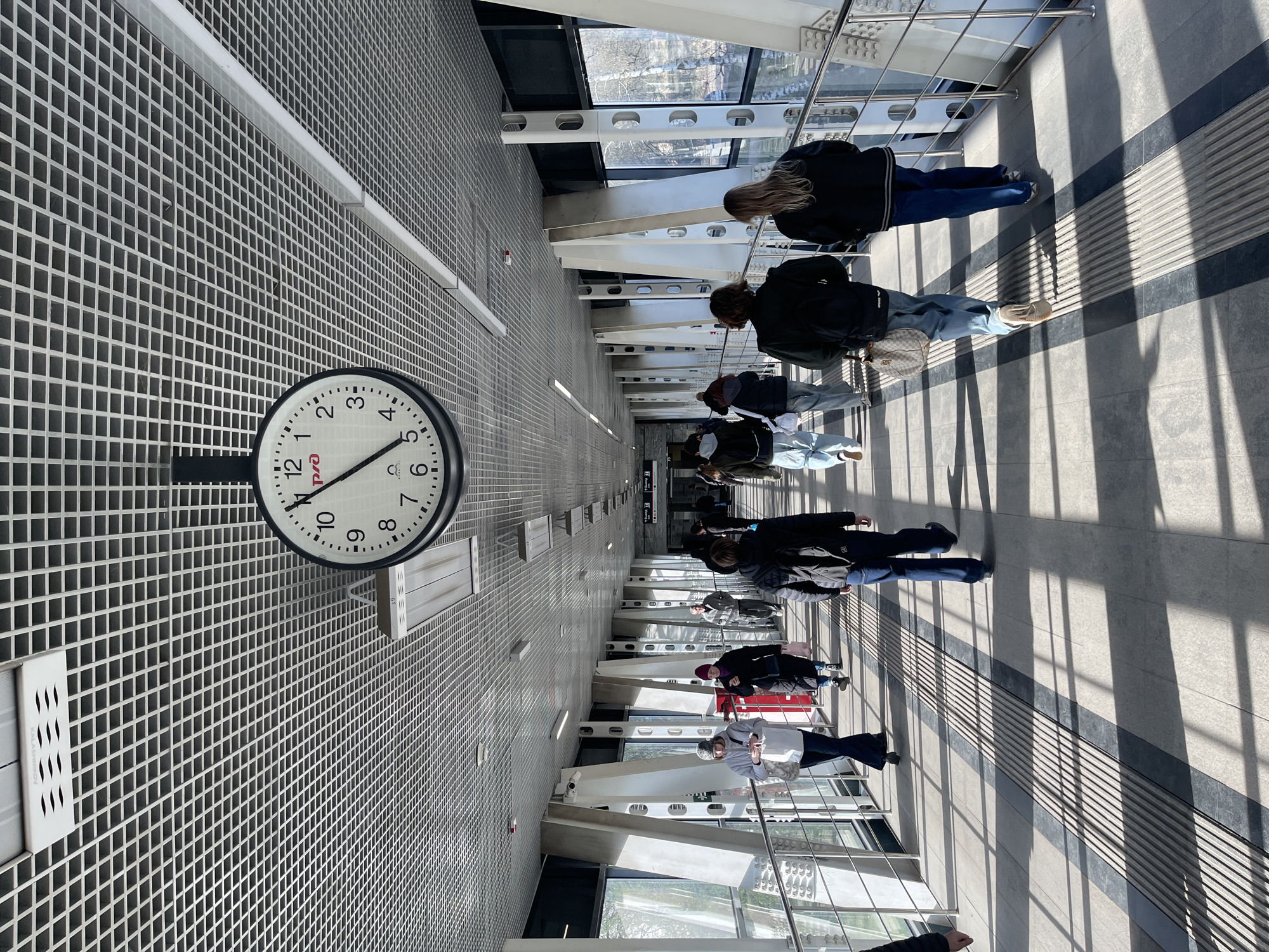 The image captures the interior of what appears to be a modern transit station in Khimki, Russia. A row of people is walking along a walkway. The walkway features tactile paving intended for visually impaired persons. Overhead structural supports with integrated window sections allow natural light to illuminate the walkway. The walls are clad in a tiled grid pattern. A clock is mounted on the wall. The time on the clock is 4:32.