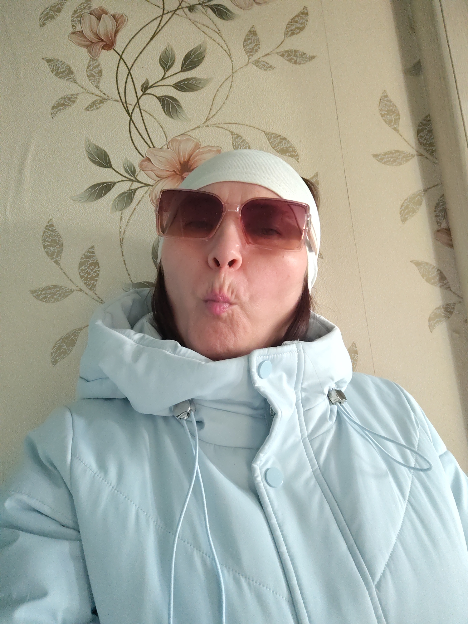 A woman is featured in a close-up, chest-up shot, appearing to take a selfie indoors. She is wearing a light blue puffer jacket with a visible hood and drawstrings, and a light-colored button closure. A white or off-white headband is worn on her head. Her face is mostly obscured by large, rectangular sunglasses with pink-tinted lenses and clear frames. She is making a playful "kissing face" or pout with her lips.
The background shows patterned wallpaper with a floral design, featuring light brown/beige flowers and green/grey leaves on a cream-colored base. A light-colored curtain or drape is visible on the right side of the frame. The setting appears to be an indoor room, likely in a residence. The puffer jacket suggests a preference for warmth. No text is visible in the image. This media file is associated with the location Сусады-Эбалак, Russia.