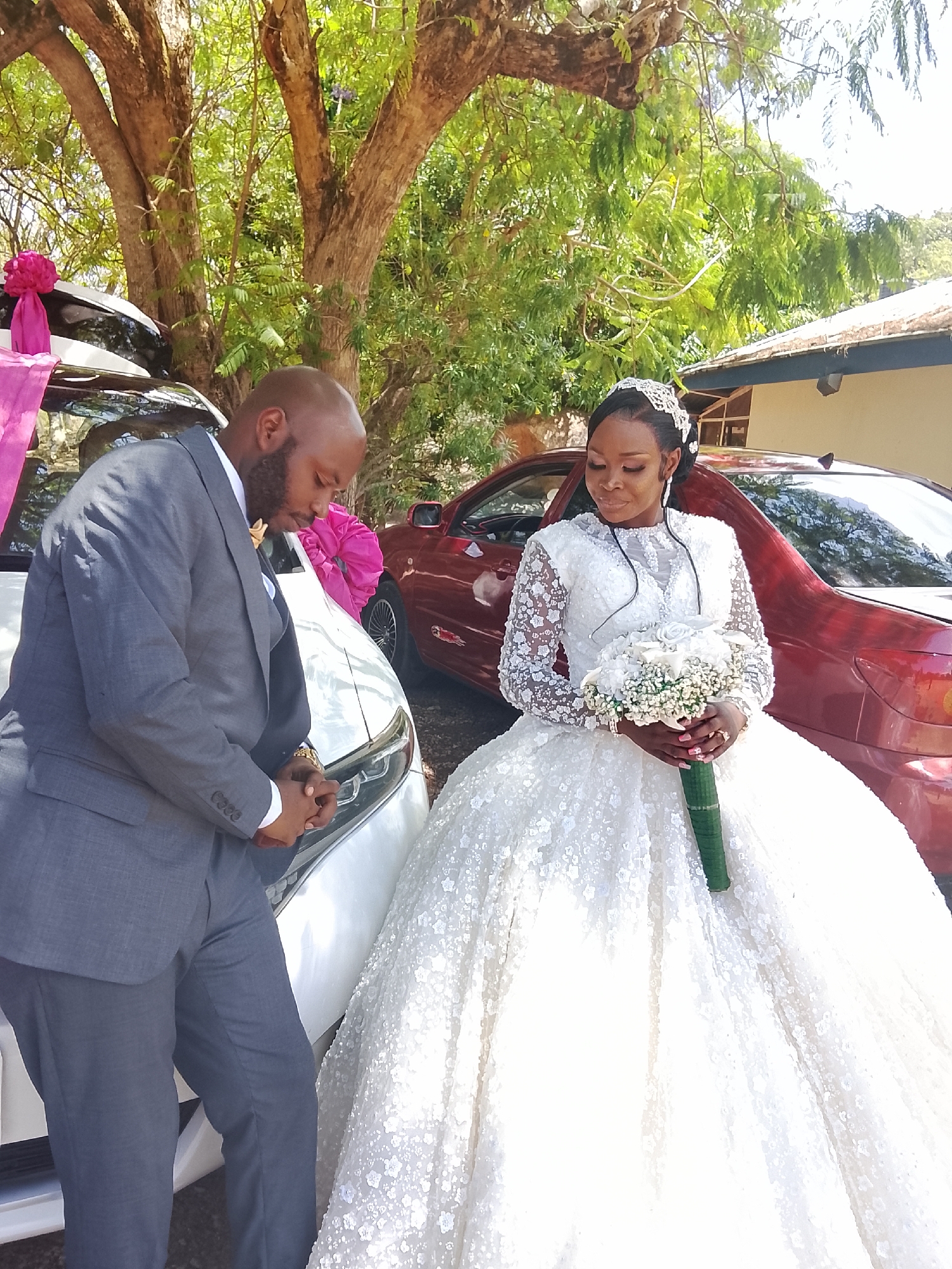 In Dutsen Kato, Nigeria, a newly married couple poses for a photograph outdoors. The groom, a bald Black man in a grey suit, stands beside a white car decorated with a pink bow. The bride, a Black woman in an elaborate white wedding gown with long, lace sleeves and a tiara, holds a bouquet of white flowers. A red car is parked behind the bride. The scene is surrounded by trees.