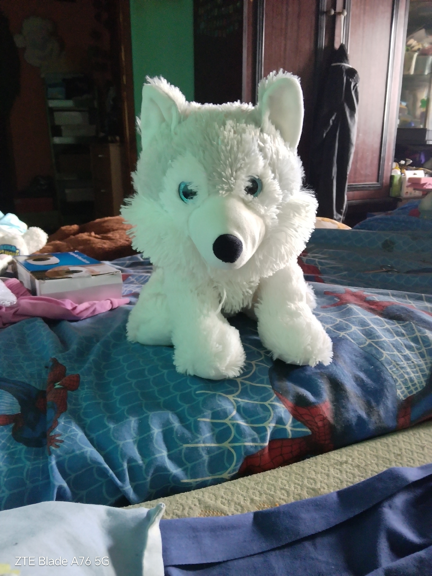 A plush white wolf toy with blue eyes sits on a bed covered with a blue comforter featuring Spider-Man. The bed is in a room with wooden furniture and a green wall. A brown blanket is visible behind the toy. The scene appears to be in a domestic setting, with no specific landmarks indicating a location in Stara Zagora, Bulgaria.