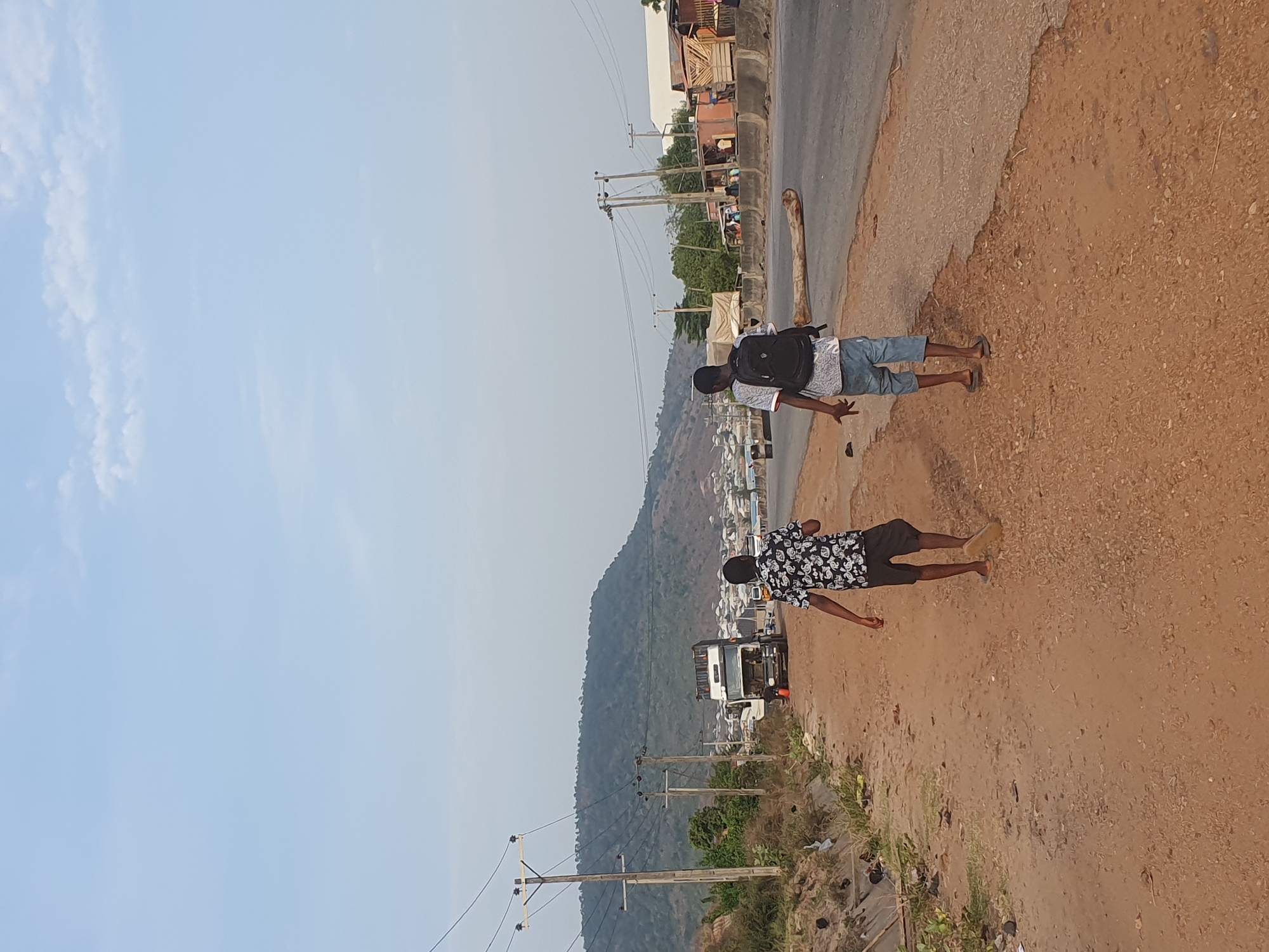 The image depicts an outdoor scene near a road in Lokoja, Nigeria. Two men are standing on the shoulder of the road, which is a dirt and gravel mixture. One man wears a light-colored patterned shirt, denim shorts, sandals, and a backpack. He is closer to the paved part of the road. The second man, closer to the foreground, is wearing a dark shirt with a floral pattern and dark shorts with sandals. A paved road runs alongside the dirt shoulder, with buildings and structures visible on the other side. Power lines run parallel to the road. In the background, a hill or mountain is visible, with buildings and a town extending down its slopes. The sky is light blue with some wispy clouds.