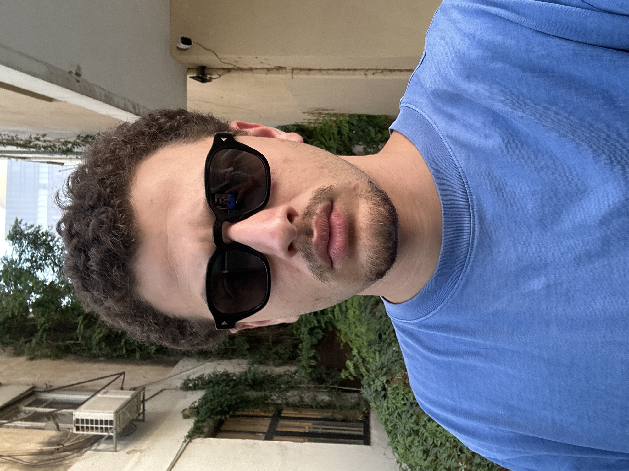 The image is a selfie of a man with curly dark hair, sunglasses, and a short beard, wearing a light blue t-shirt. He is outdoors, with a building as the backdrop, possibly in Tel Aviv, Israel, given the presence of buildings with visible air conditioning units and vegetation often found in urban environments there. The building is old, with overgrown plants and window gratings. The photo is taken at a slightly rotated angle. A security camera is visible on the building in the background.