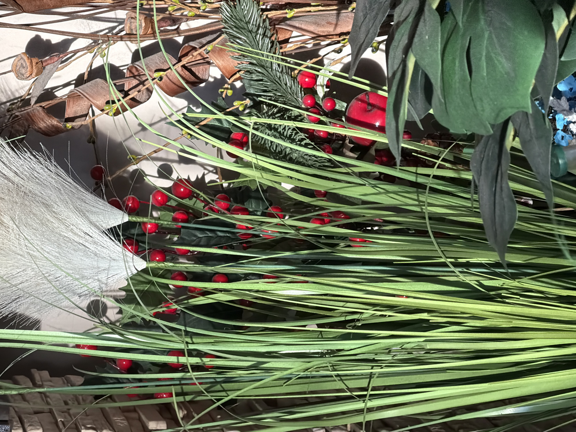 The image shows a collection of decorative holiday items. There are green artificial grasses, red berries, brown coiled twigs, a white feathered decoration, and dark green leaves. A small section of pine needles and a red apple-shaped ornament are also visible. The background is light-colored. The location and city are unknown.