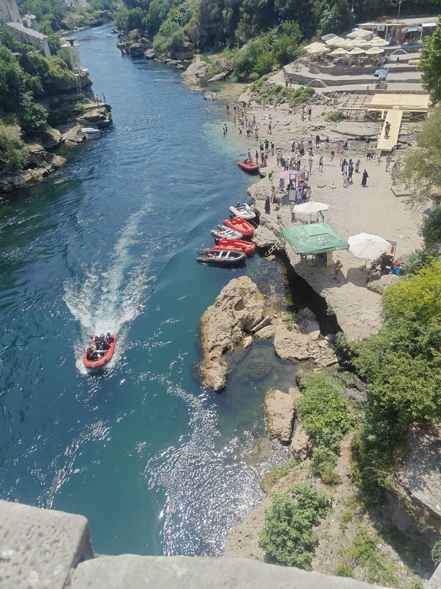 The image shows a wide river flowing through a city, with a beach area populated by many people on the right bank. The river is deep blue and appears to be flowing swiftly, with a visible wake from a small red boat carrying several people. Several other small boats are docked on the left bank near a rocky outcrop. On the right bank, a sandy beach is lined with umbrellas and seating areas, suggesting a recreational spot. People are seen swimming in the river, sunbathing on the beach, and milling around the various amenities. Green trees and vegetation frame the scene, indicating a warm, sunny day. This scene is likely the Neretva River in Mostar, Bosnia and Herzegovina, near the Stari Most (Old Bridge) area, known for its natural beauty and recreational activities.
