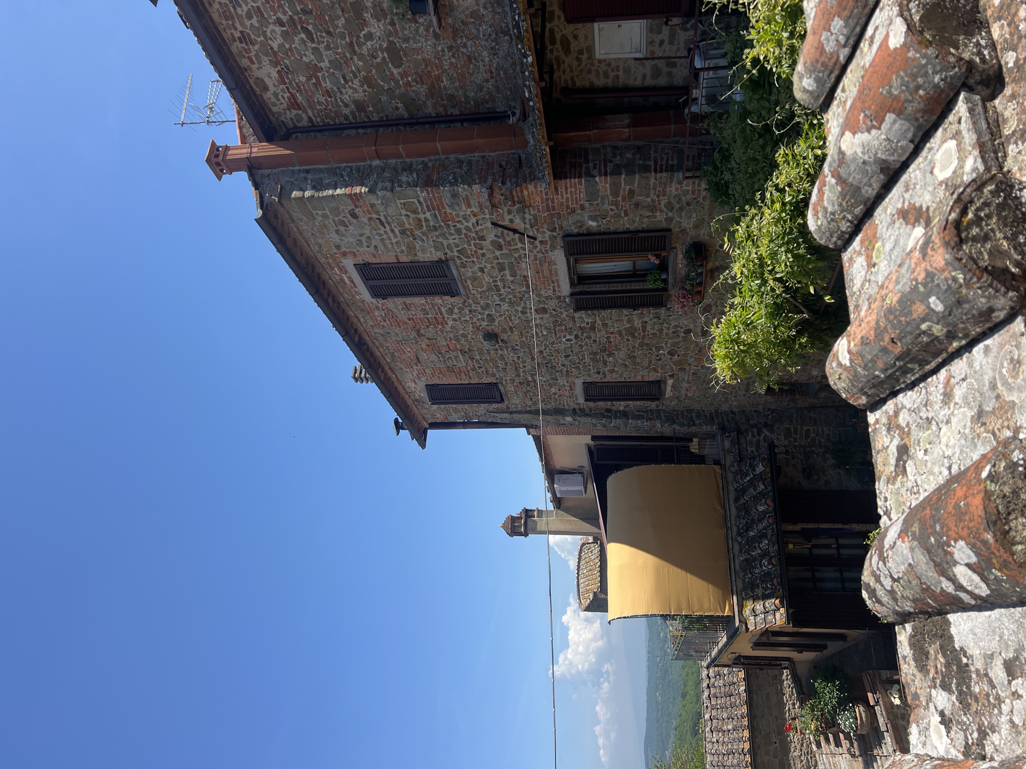 Captured from a slightly elevated perspective, the image showcases architecture within the town of Sinalunga, Italy, set against a clear blue sky. A multi-storied building is constructed of stone with dark-shuttered windows. Patches of greenery are visible between some windows. A light-colored awning is positioned on a lower level. The image's foreground features aged, lichen-covered brickwork, likely part of the observation point. A landscape with mountains is visible.