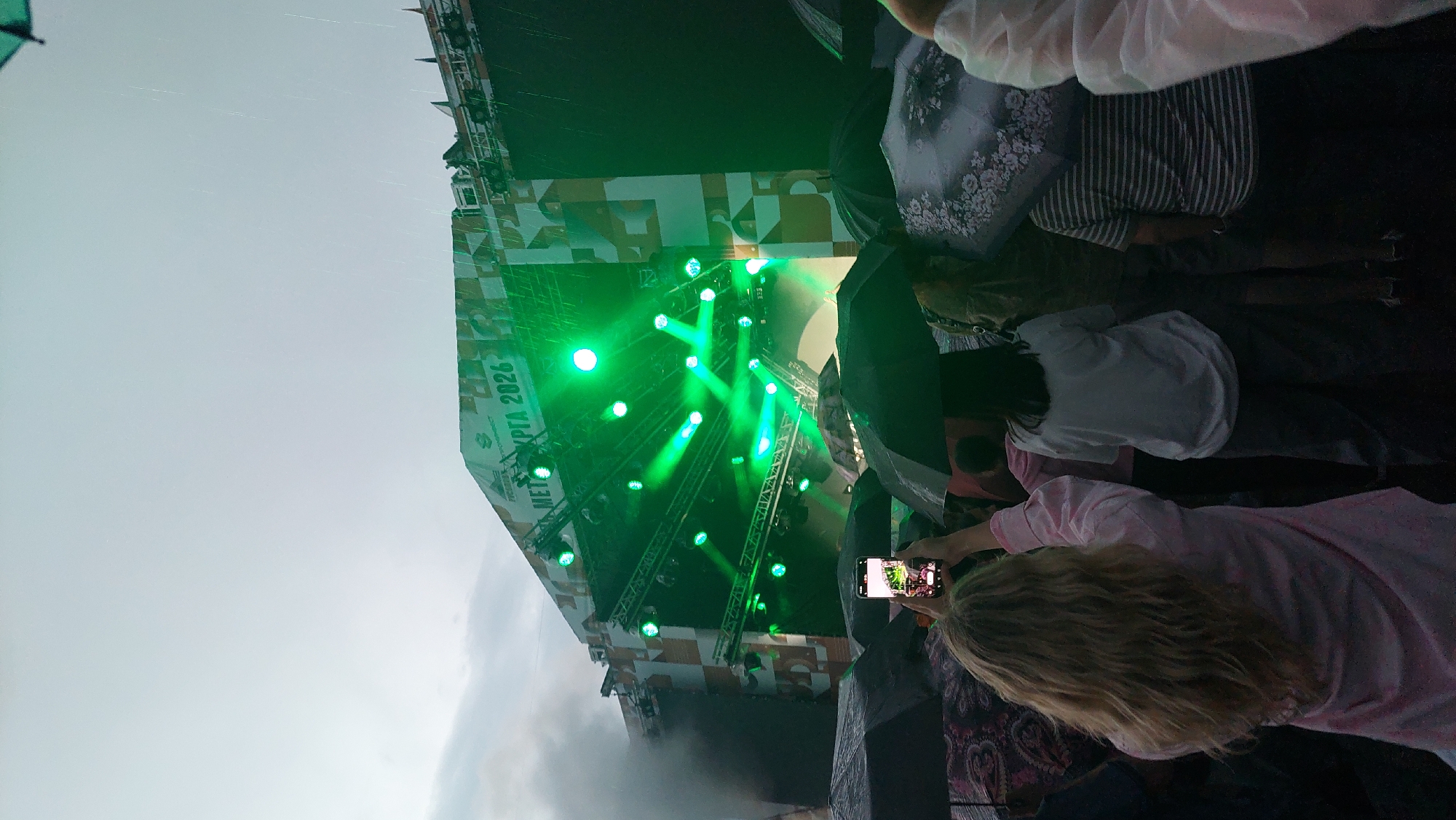 An outdoor concert is taking place in Solnechnyy, Russia. The stage is lit with green lights, and a banner reads "METKPGA 2026". It is raining, and the audience is holding umbrellas. A woman in the foreground is taking a picture of the stage with her phone. The image captures a moment of shared experience, with the crowd united by music and the shared challenge of the weather.