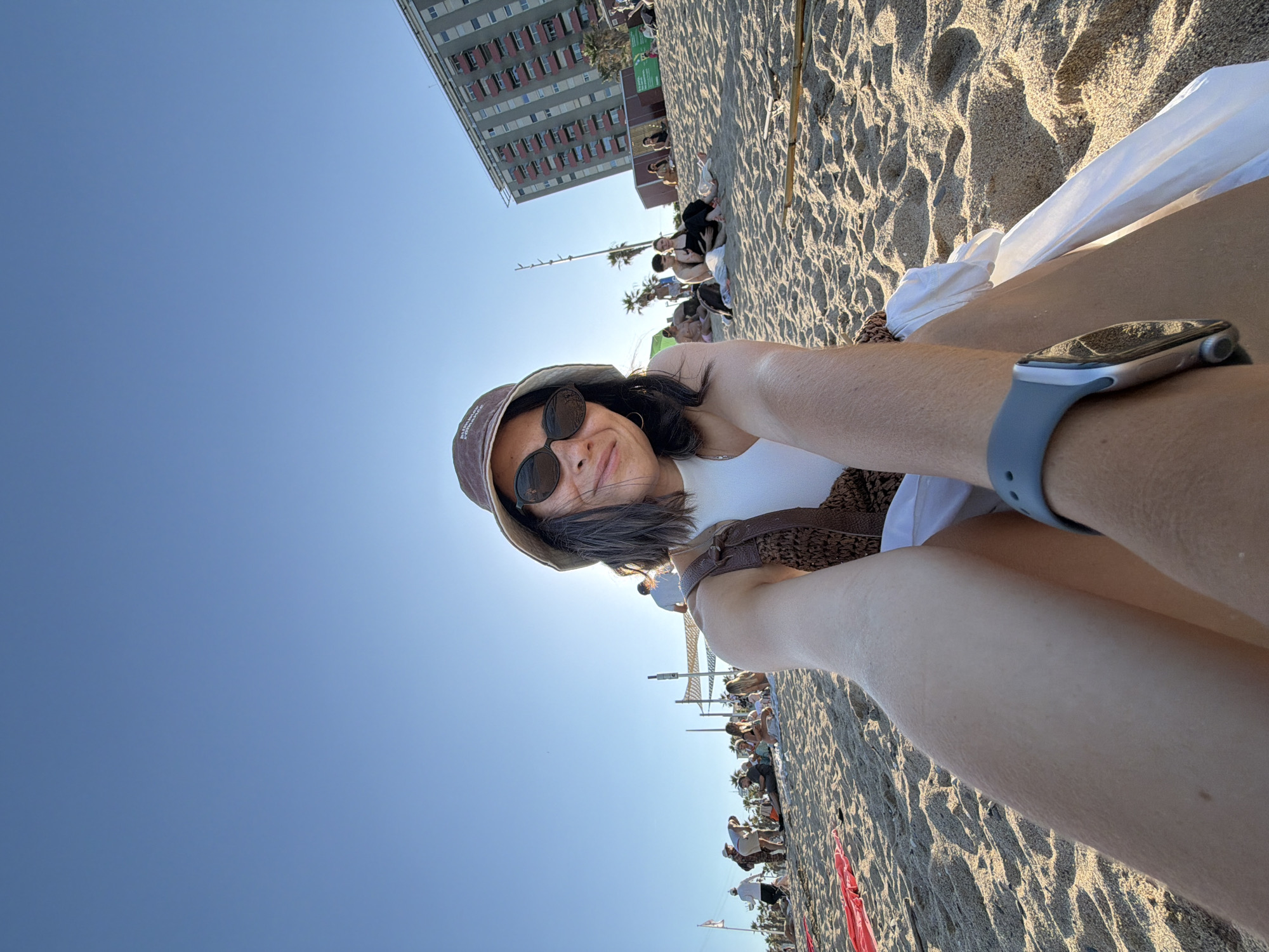 The image shows a woman wearing a bucket hat and sunglasses, sitting on a sandy beach in Barcelona, Spain. She is taking a selfie, with her arm and part of her leg visible in the foreground. She is wearing a white top and a brown crocheted vest. Behind her, other beachgoers are scattered on the sand, and a tall, multi-story building with balconies can be seen in the background. The sky is clear and blue, and the sun is shining brightly. The scene appears to be a sunny day at the beach, likely in the late afternoon given the lighting and long shadows.