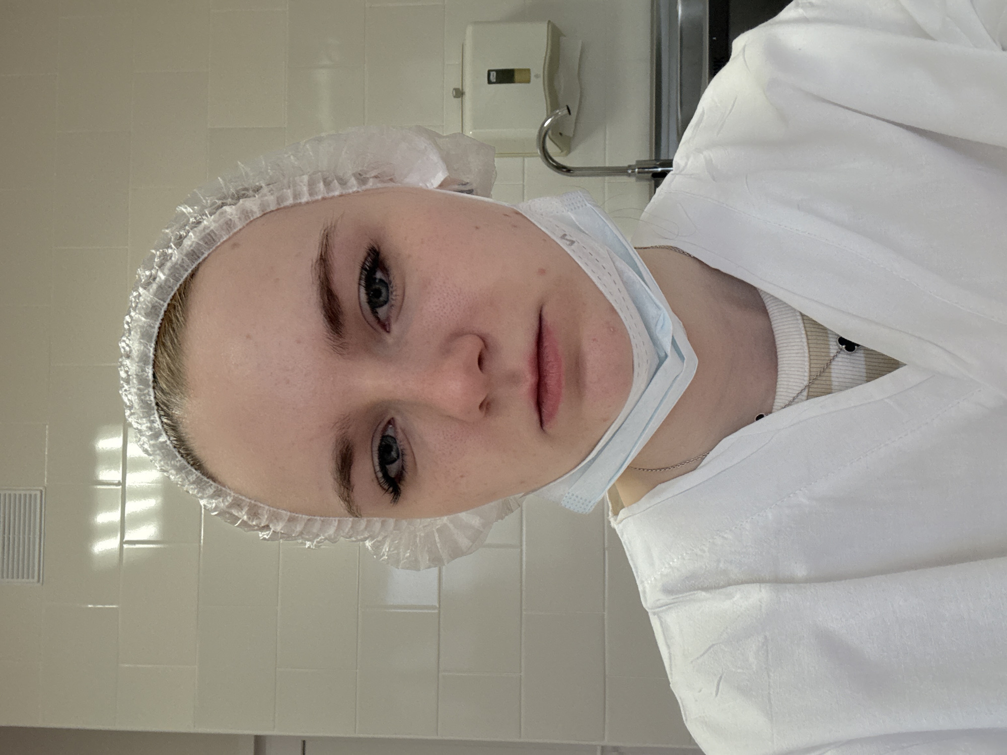 A young woman wearing a white surgical cap and a white lab coat is shown in a close-up portrait. A blue surgical mask hangs loosely around her neck. She has fair skin, blue eyes, and dark eyebrows. Her expression is neutral. The background consists of white tiled walls and a stainless steel sink with a faucet. A white dispenser is mounted on the wall behind her. The location is consistent with a medical or laboratory setting. There is no information to identify if the location is in the city of Saint Petersburg, Russia.