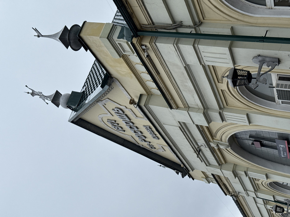 This image depicts the facade and roofline of the Rossosh railway station building in Rossosh, Russia. The building features a light yellow main color scheme with lighter, possibly off-white or light gray, decorative trim, pilasters, and cornices.
The prominent gable section of the roof, covered with dark, possibly green or black, tiled material, displays raised white lettering: "1871" at the apex, followed by "ст. РОССОШЬ" (st. ROSSOSH), and smaller text "ОВЖД" (OVZhD) beneath "РОССОШЬ." Two metallic finials adorn the roofline; the central finial on the gable peak is a stylized, multi-part figure (possibly a winged form or a locomotive representation) resting on a spherical base. To the right, a simpler, spired finial is positioned on a lower section of the roof.
Below the gable, a small, light-colored speaker or horn is mounted on the facade. Further down, two small, light-colored security cameras are affixed to the facade below a cornice on the right side. A vertical green downspout or utility conduit runs along the far right edge of the building. On the lower facade, an ornate, black, lantern-style wall sconce with a decorative bracket is attached to a light-colored pilaster. Arched window or door openings are visible; below one on the left, a red sign with white text "Выход в город" (Exit to city) and "Way out" is partially visible. An external air conditioning unit is positioned below another arched opening.
The sky is overcast and uniformly light gray, indicating a cloudy day. No individuals or active interactions are visible in the frame.