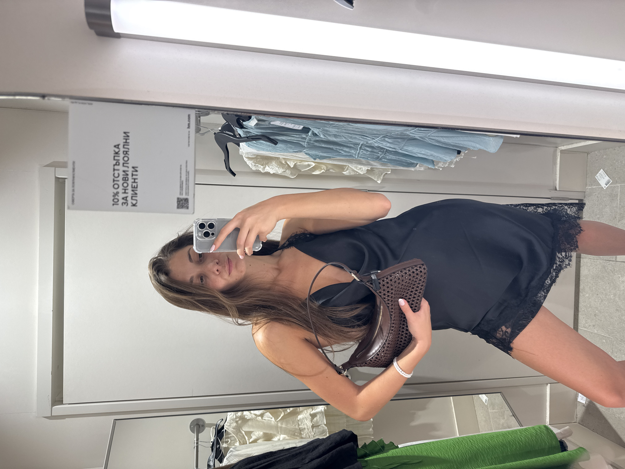 A young woman is depicted taking a mirror selfie within what appears to be a retail store environment, likely a fitting room, in Burgas, Bulgaria. The image shows her standing, holding a silver-colored smartphone in her right hand to capture her reflection, while her left hand holds a stylish brown woven handbag. She is wearing a black slip dress adorned with delicate black lace trim at the neckline and hem. Her long, dark brown hair falls over her shoulders, and she presents a neutral, pleasant expression, looking directly into the camera. A white beaded bracelet is visible on her left wrist, complementing her light pink nail polish.
The background, reflected in the mirror, features clothing racks displaying various garments, with light blue, white, and green fabrics visible. The setting is characterized by white walls, light-colored flooring, and bright overhead fluorescent lighting, typical of commercial retail spaces.
A white sign, prominently reflected in the upper left portion of the mirror, provides specific location cues. The text on the sign, written in Cyrillic, states "10% ОТСТЪПКА ЗА НОВИ ЛОЯЛНИ КЛИЕНТИ," translating to "10% DISCOUNT FOR NEW LOYAL CUSTOMERS." Below this, the website "hm.com" and a QR code are visible, strongly indicating the store is H&M. A partial view of another mirror is seen at the bottom of the main mirror. The scene captures a moment of personal style and shopping.