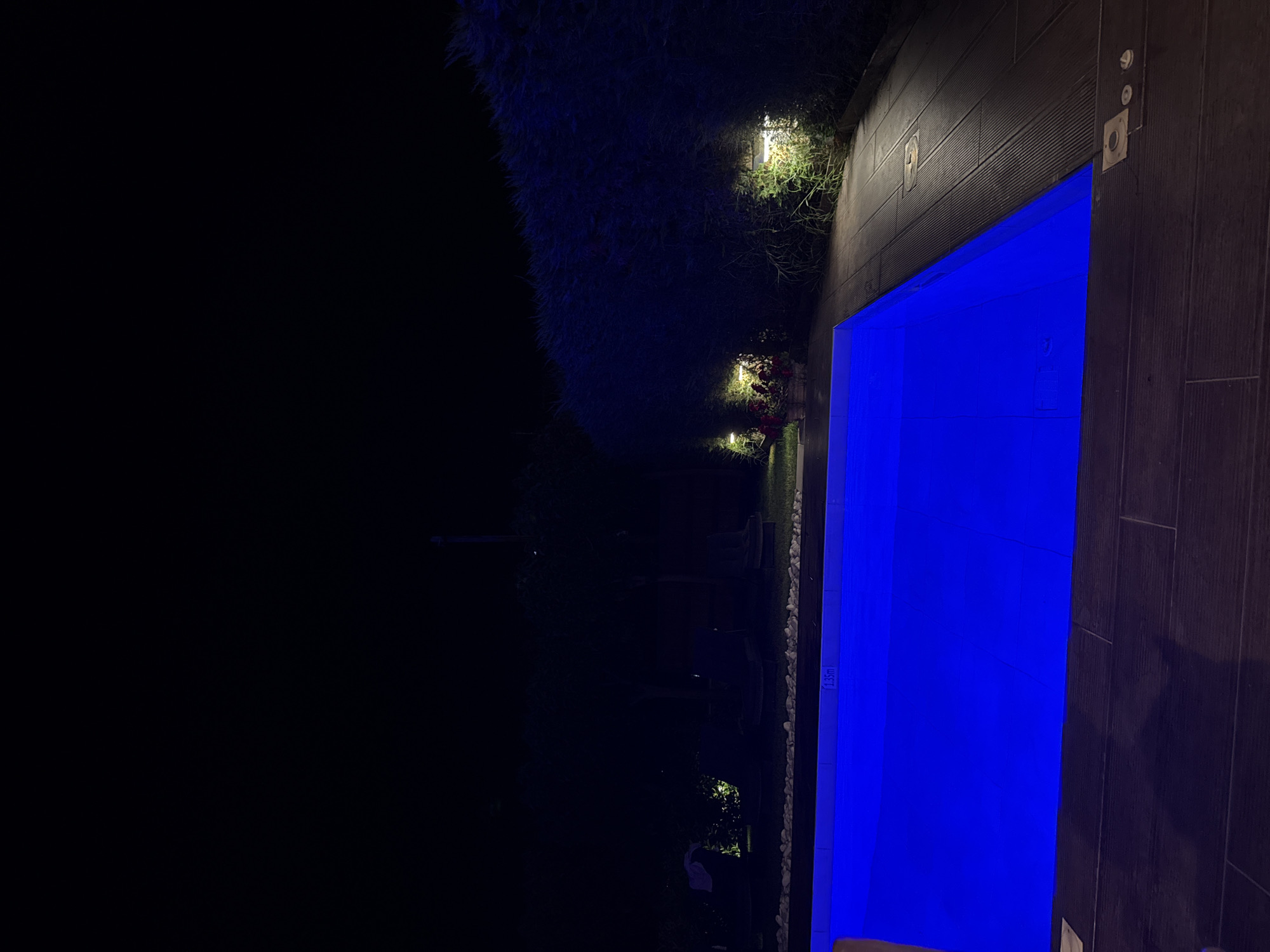 This media file is a photograph taken at night, showcasing a swimming pool illuminated with a vibrant blue light. The pool is situated next to a dark wooden deck, with some decorative stones lining the edge of the water. To the left of the pool, there is a dark area that appears to be a garden or outdoor space, with some foliage and multiple wall-mounted lights casting warm yellow glows. One of these lights is visible illuminating a patch of greenery and some red flowers. The overall atmosphere is serene and inviting, suggesting a tranquil evening setting. The image is oriented sideways. A visible text label on the pool's edge reads "1.35m". The location context is Pallini Municipal Unit, Greece.
