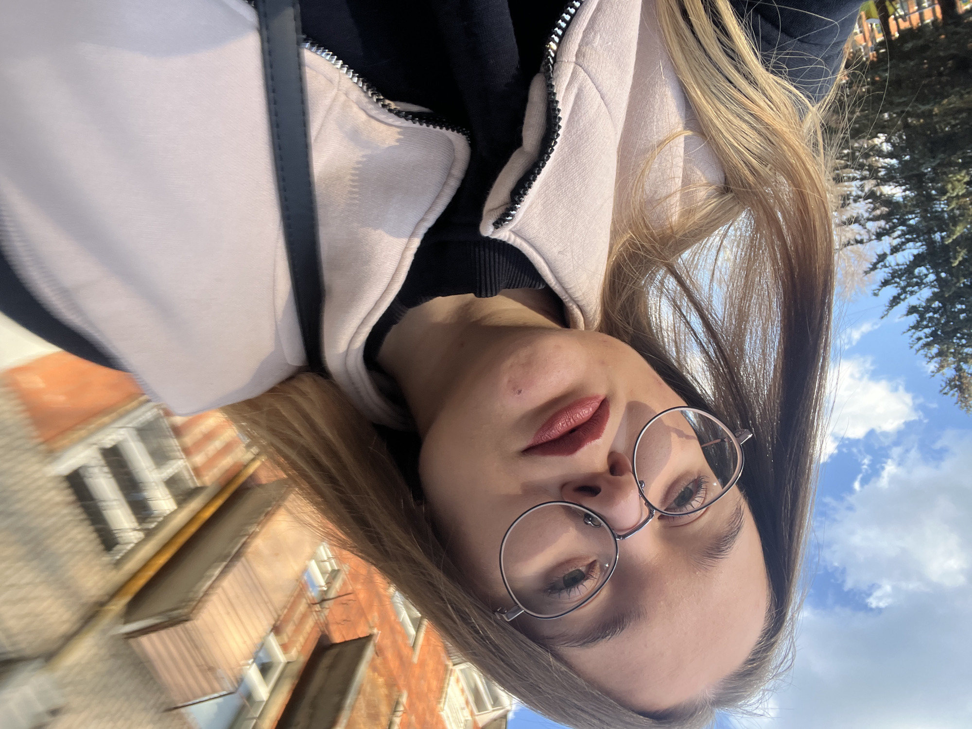 A woman with long blonde hair and glasses is shown in a selfie outdoors. She is wearing a light pink zip-up jacket over a black shirt. In the background, there is a brick building on the left and trees and a cloudy blue sky on the right. The photo was taken in Pushchino, Russia.
