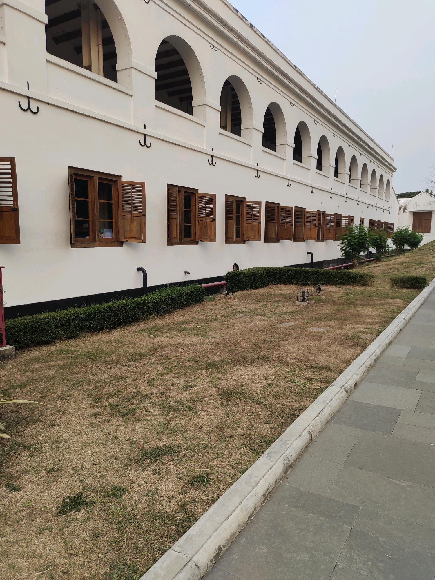 The image depicts the exterior of a white colonial-style building with a long arched facade. The building has numerous wooden-shuttered windows on the ground floor and arched openings on the upper level. The facade is adorned with black metal hooks spaced along the upper section. In front of the building, there is a well-maintained hedge bordering a lawn with dry, brown grass. A paved walkway runs along the right side of the scene, separated from the lawn by a concrete curb. The sky is overcast. The building appears to be part of Fort Van den Bosch in Ngawi, Indonesia, a historical site from the Dutch colonial era. There are no people visible in the image.