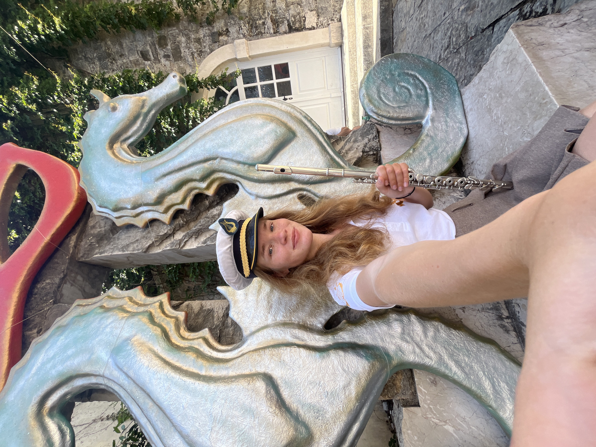 A young person wearing a white captain's hat and a white shirt poses with a flute in front of a large, decorative sculpture of sea creatures. The sculpture features two seahorses rendered in a silvery-green color with golden accents. The individual is lying on the sculpture, holding the flute towards the camera as if playing it.
Behind the sculpture, there is a light-colored stone wall covered with green vegetation. A white door with windows is visible in the background. The scene appears to be outdoors, in a sunny setting. The location is in the city of Budva, Montenegro.