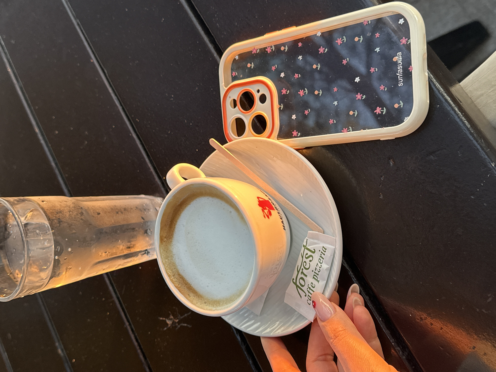 The scene is a close-up shot on a dark, wooden, slatted table. A phone case with a cream-colored border and a black background decorated with pink and white floral patterns, labeled "sunfasuota," rests on the table. Next to it is a white cup filled with a latte, placed on a matching saucer with a small wooden stirrer and a packet of sugar labeled "forest caffe pizzeria." To the left of the cup is a partially filled clear glass of water. A person's hand, with a light skin tone and manicured nails (one with a small strawberry design), is holding the sugar packet.
This scene, featuring "forest caffe pizzeria," indicates the image was taken in or near Šušanj, Montenegro, as this is the location of the establishment.