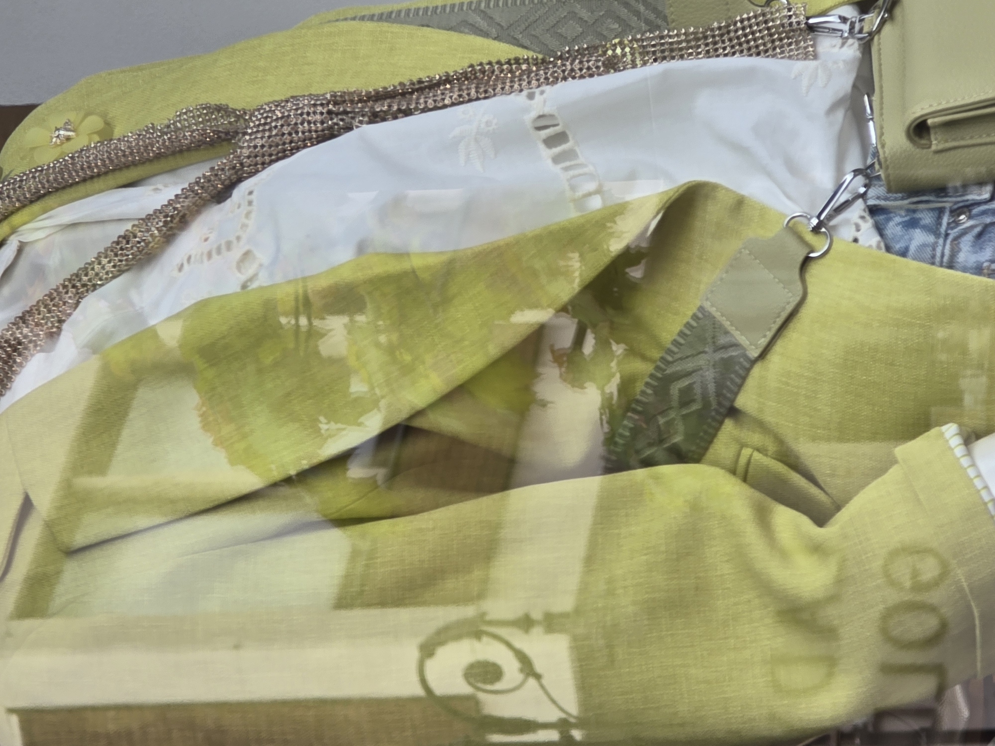 A close-up, semi-obstructed view of garments arranged in a display case. The garments include a yellow-green jacket, a white blouse with lace details, a metallic belt, and a small yellow-green purse. The lower portion of a pair of jeans is also visible. The scene is set in the city of Unknown, Unknown, but the exact location within the city cannot be determined from the image. The photograph is taken through a glass surface, causing some reflections and making the objects appear slightly distorted.
