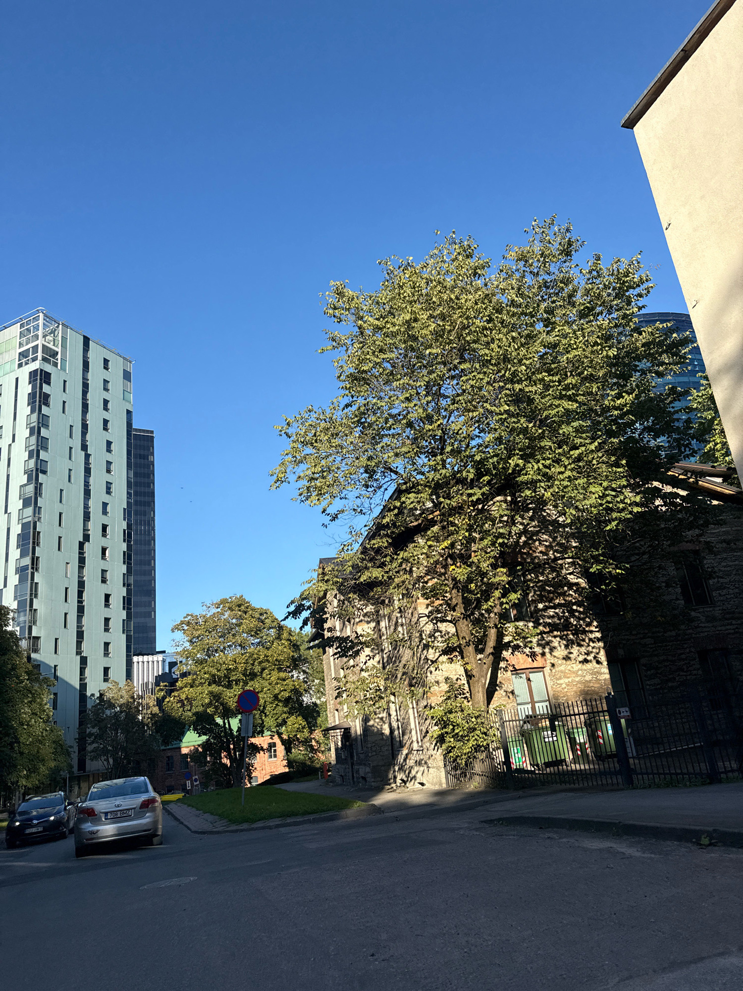 The image depicts a street scene in Tallinn, Estonia, with a focus on architecture and urban landscaping under a clear blue sky. Dominating the left side of the frame is a tall, modern, mint-green skyscraper with numerous windows and a glass-paneled top. To its right, a darker, equally tall building with a glass facade is partially visible. In the foreground, a paved road curves gently, with two cars parked on the left side of the road. The closest car is a silver sedan with license plate 708 BMZ, followed by a dark blue hatchback.
In the middle ground, a mature tree with lush green foliage casts a shadow across a stone building. This building appears older, constructed from rough-hewn stones, and features dark-paned windows. A black metal fence encloses a small area with green trash receptacles behind it. To the right of the stone building, a light-colored, modern structure with a sloped roof juts into the frame from the right edge, partially obscuring the view of the sky.
Behind the stone building and to the left, a smaller, red-brick building with green-tiled roofs is visible, surrounded by greenery. A circular sign with a red border and a blue center, indicating a no-entry zone, is placed on a pole near the grass area in front of the brick building. The overall impression is a blend of modern and historic architecture within a city environment.