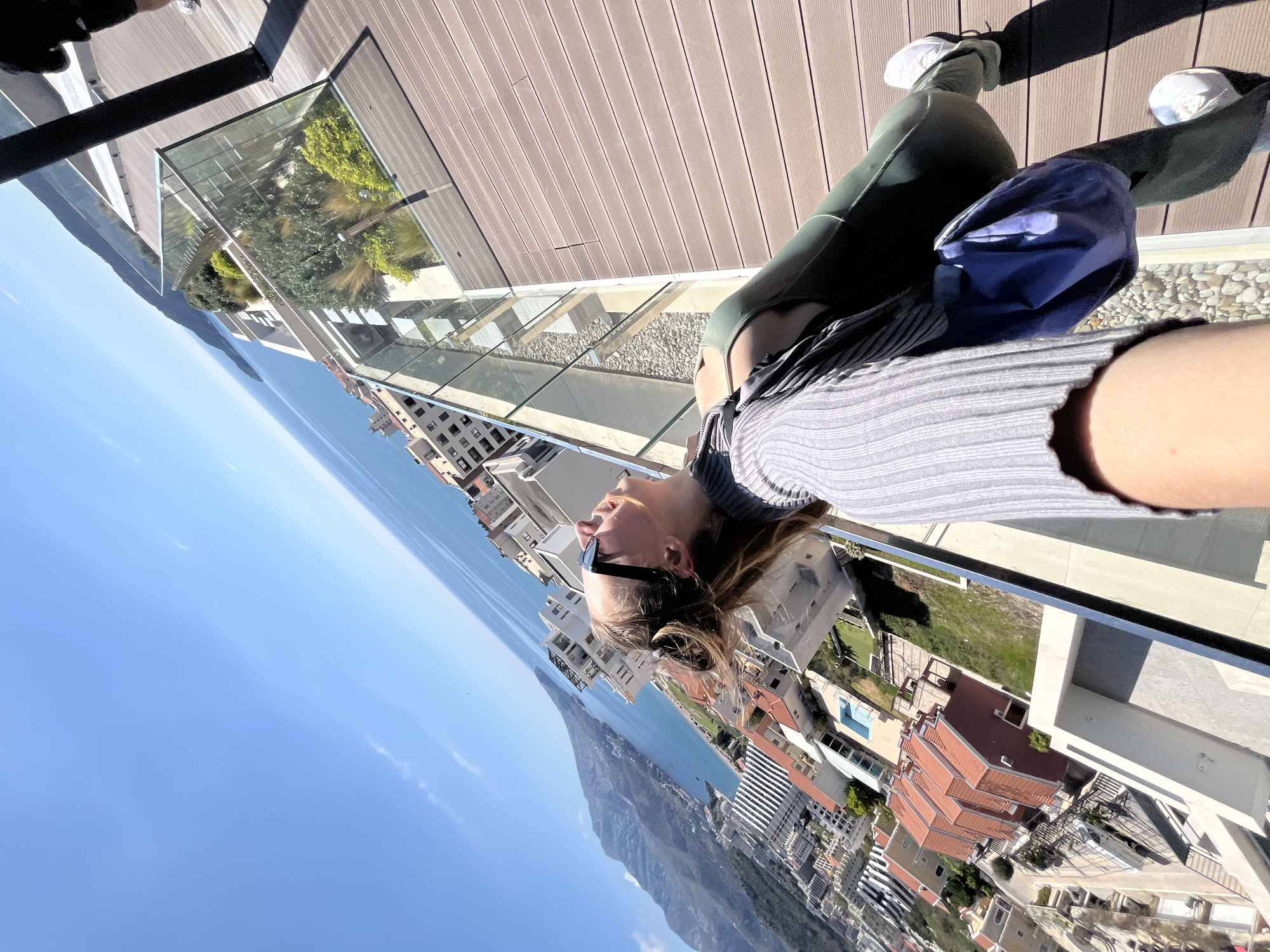 A medium shot depicts a woman in a gray long-sleeved shirt, green leggings, and white sneakers leaning on a balcony rail. She is wearing sunglasses and looking up, with buildings and a mountain visible in the background. The sky is blue with a few clouds. The location appears to be Boreti, Montenegro, as the architecture and landscape are consistent with this city.