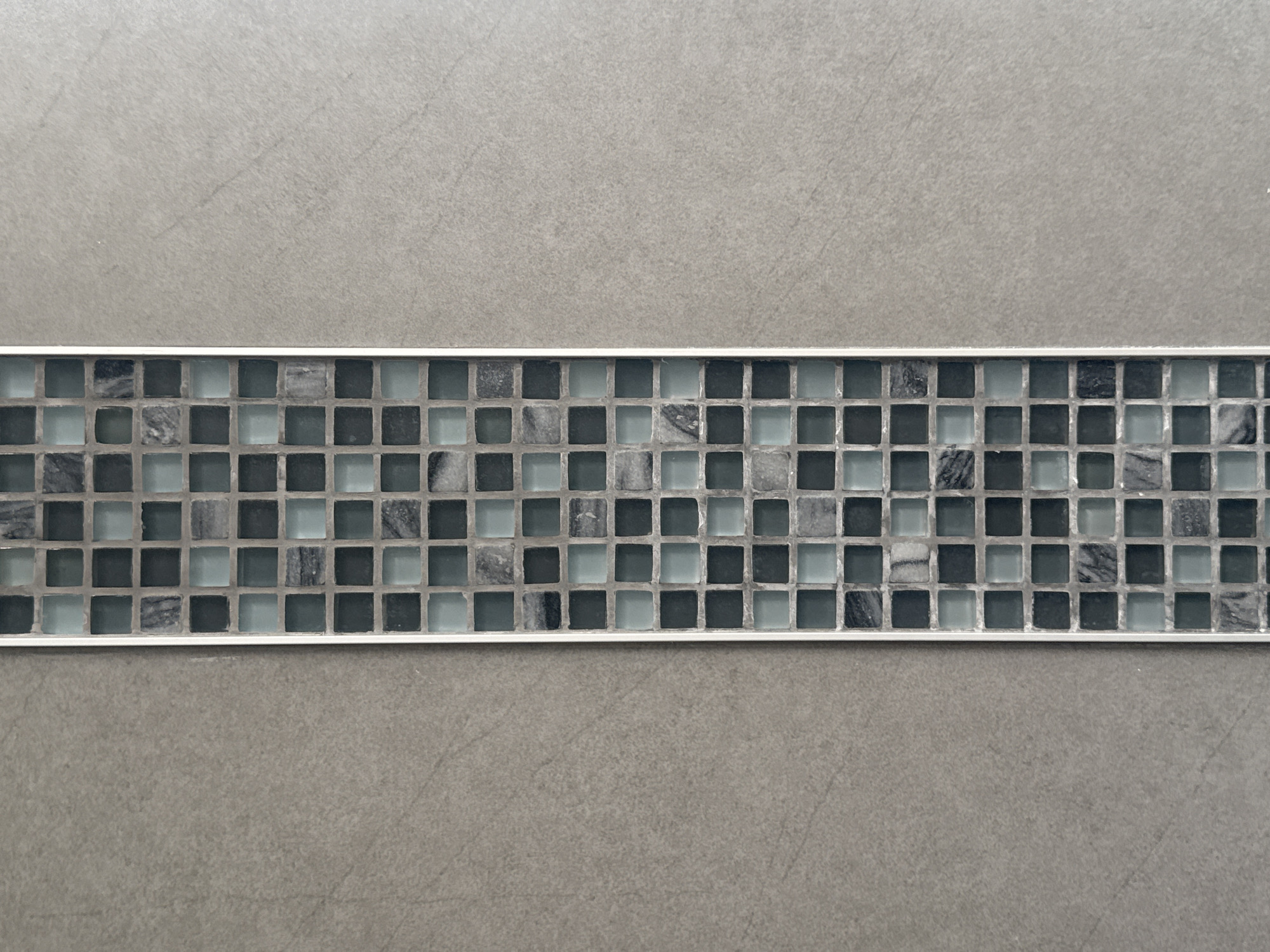 A close-up shot presents a wall composed of a gray, possibly concrete-like material. Across the center, there is a horizontal strip of small, square tiles of varying shades of gray and blue, framed by a thin, metallic border.