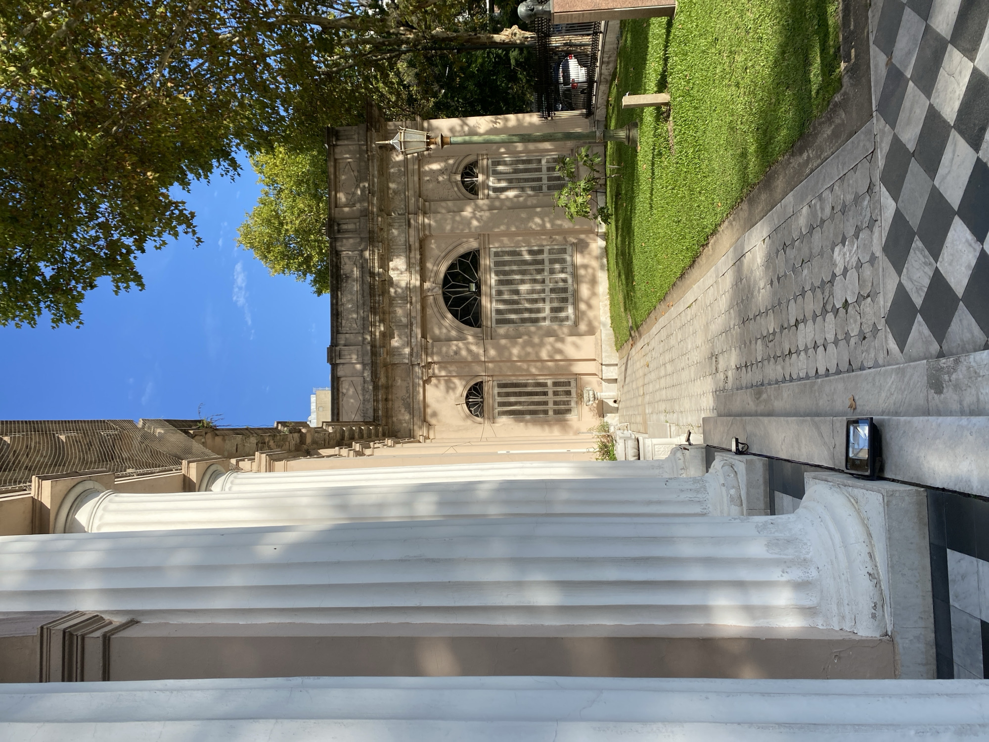 The image shows the facade of the Museo de Arte Hispanoamericano Isaac Fernández Blanco in Buenos Aires, Argentina. The building is a two-story, light-colored structure with arched windows and decorative shutters. A row of white classical columns lines the front of the building, casting shadows on the staircase. A sidewalk with black and white checkered tiles runs alongside the building, separating it from a patch of green grass. Trees are visible above the building, and a clear blue sky is above the trees. A lamppost is visible further down the sidewalk, beyond the building. A small section of a black metal fence, beyond the grass patch, is also visible.