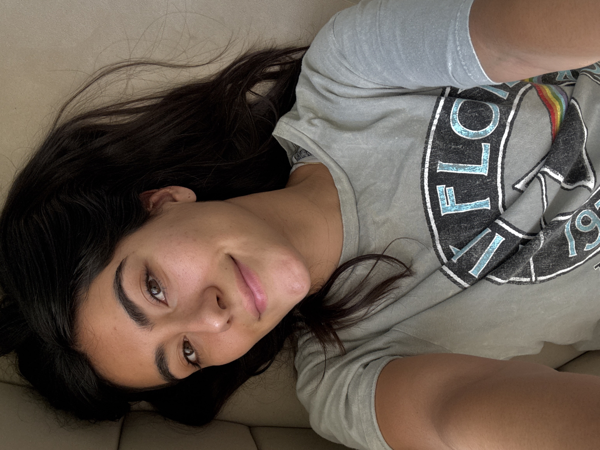 A young woman with dark hair is lying on a light-colored sofa, facing the camera. She is wearing a grey t-shirt with a circular design featuring the word "FLOYD." Her hair is spread out behind her head on the sofa. The image appears to be taken indoors, under natural lighting. There is no indication of the location being Puebla, Mexico, or any information about the environment beyond the immediate surroundings of the subject.