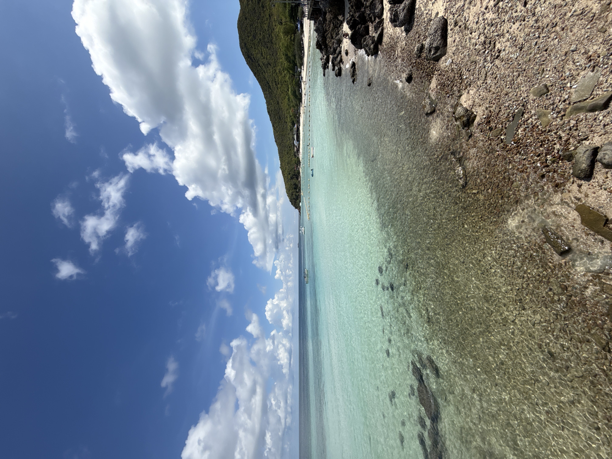 The image shows a beach scene in Pattaya City, Thailand. A coastline dominated by a green, vegetation-covered hill extends along the upper left. Clear turquoise water borders the beach. Several boats are visible in the distance. The sky above is a vibrant blue, dotted with white clouds. The beach itself consists of sand and pebbles, merging into larger rocks at the right edge of the frame. The orientation of the photo is sideways.