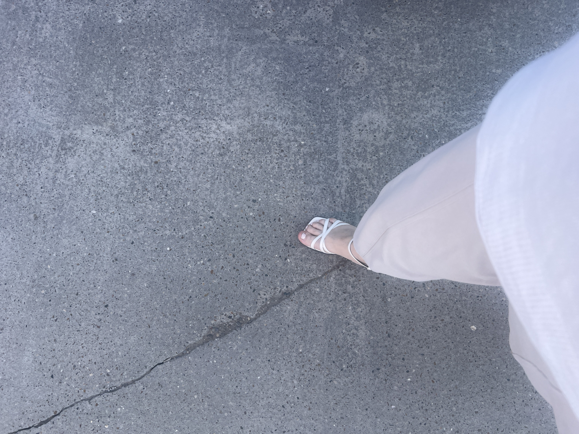 The image shows a person's right foot wearing a white strappy sandal on a cracked gray concrete surface. The person is wearing beige pants and a white blouse, with a portion of the blouse visible in the upper right of the frame. The concrete surface is textured with small pebbles and a crack runs diagonally across the bottom left corner of the image, passing near the foot. The image appears to be taken from a high angle. There is no specific evidence to indicate the location is in Chaikovsky, Russia.