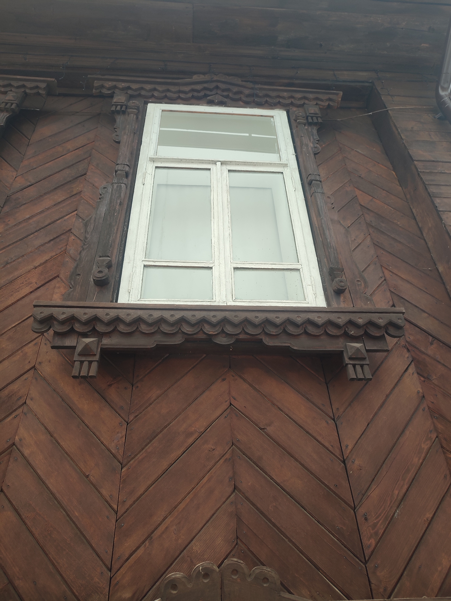 A low-angle medium shot features a window on a building in Sarapul, Russia. The window is a white-framed double-hung unit with six panes. The building's exterior is covered in brown wooden siding arranged in a herringbone pattern. Ornate dark wood carvings frame the window, including a scalloped decorative piece above the window sill and sculpted supports. Fasteners, likely nails or screws, are visible throughout the wooden siding. The sky is overcast and gray.