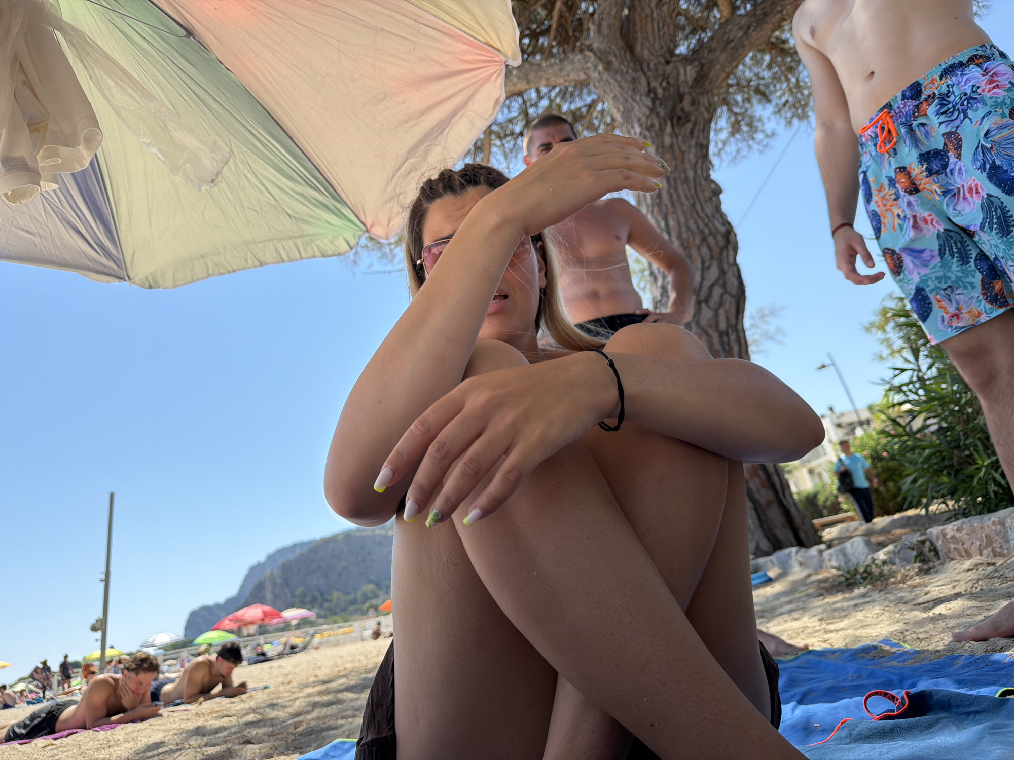 The image depicts a sunny day at a beach, possibly Mondello Beach in Palermo, Italy, given the mountainous backdrop. In the foreground, a woman is seated on a blue towel, partially obscured by a large, colorful umbrella overhead. She is wearing sunglasses and has her hand raised towards her face, her nails painted with a yellow and white design. To her right, a man in patterned swim shorts stands, his torso bare. Behind them, a second man, also shirtless, is visible, looking towards the viewer. The beach itself is sandy, with other beachgoers and umbrellas in the background, and lush greenery and buildings further in the distance. The sky is a clear blue.
