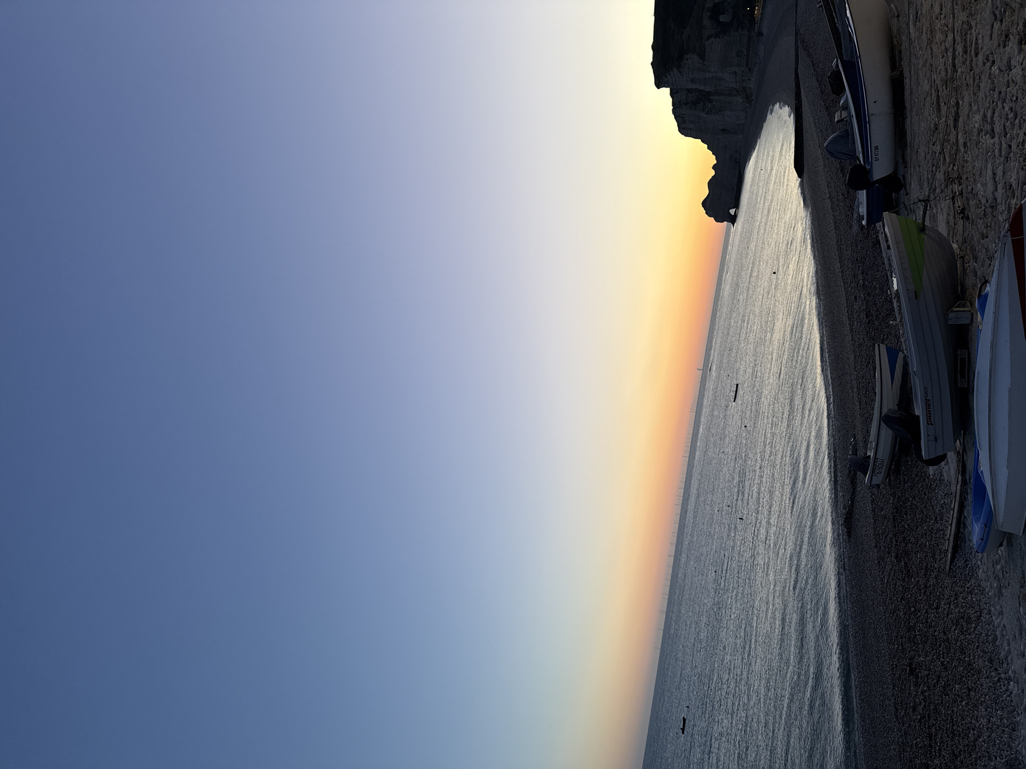 The image depicts a serene coastal scene at dusk in Étretat, France. The sky transitions from a deep blue at the top to a gradient of soft orange and yellow near the horizon, indicating the setting sun. The sea is calm, with gentle waves lapping against a pebble beach. In the distance, a line of wind turbines stretches across the water, and the iconic cliffs of Étretat, including the natural arch, are silhouetted against the vibrant sky. Several small boats are pulled up onto the beach, suggesting a fishing or recreational area. The overall atmosphere is peaceful and picturesque, capturing the beauty of the Normandy coast at the end of the day.