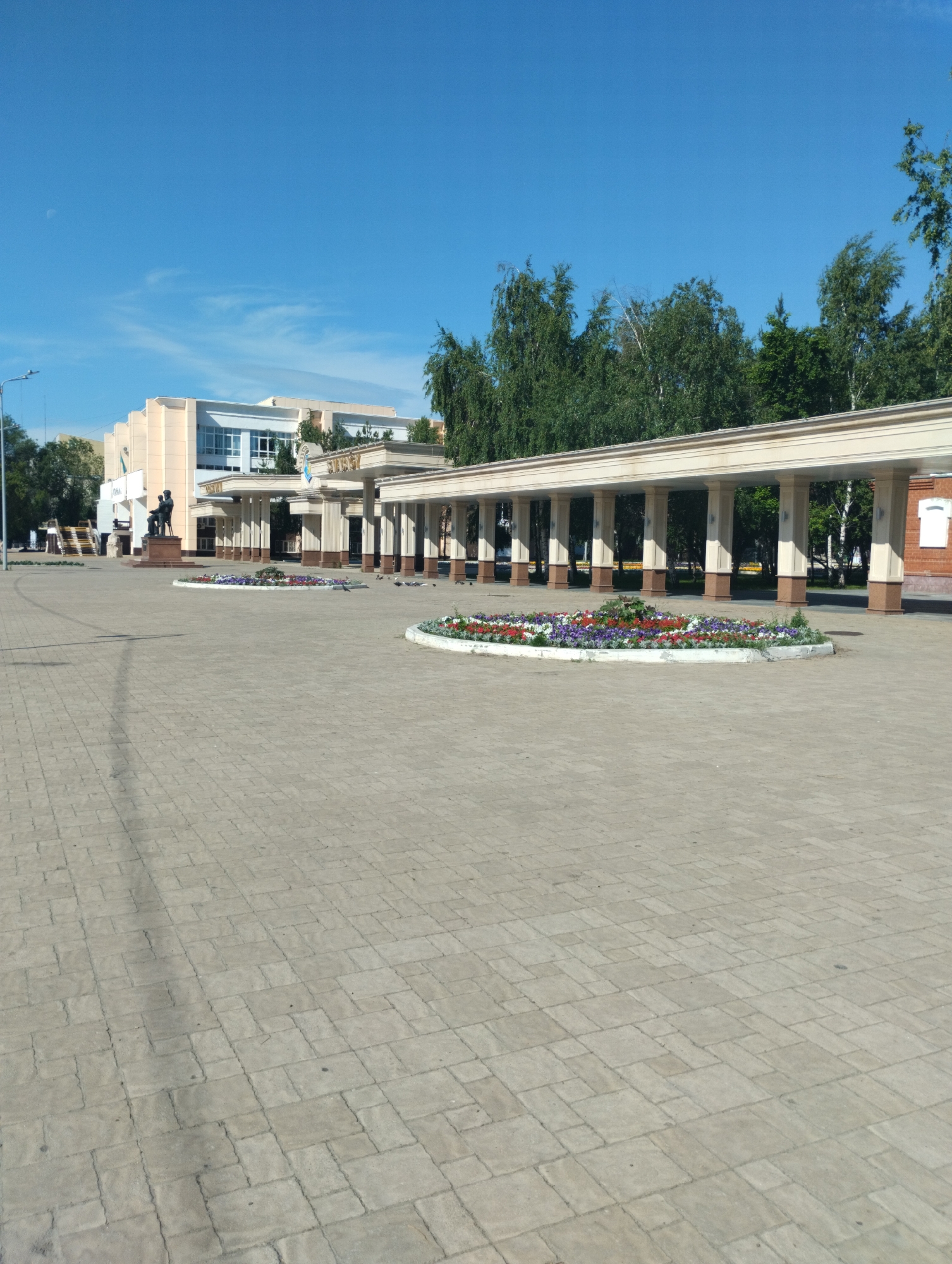 The image displays an outdoor public square situated in Kostanay, Kazakhstan, specifically identifying as the plaza fronting the Kostanay Regional Philharmonic named after E. Umirzakov. The scene is illuminated by daylight under a clear blue sky, featuring a faint crescent moon in the upper-left corner and sparse, wispy clouds.
The immediate foreground is characterized by an extensive paved surface composed of light grey interlocking concrete pavers of varying rectangular and square shapes. Two circular, raised flower beds are centrally positioned in the midground, containing an assortment of red, purple, and white flowering plants. Scattered across the paved area, particularly around the flower beds and beneath the colonnade, are multiple pigeons.
On the left side of the frame, a prominent building with a multi-story structure is visible. Its facade is a combination of light peach-toned panels and white architectural details, featuring large, multi-paned windows on its upper levels. In front of this building, a dark-colored statue of a seated human figure rests on a reddish-brown pedestal. This statue depicts Ahmet Baitursynov.
Extending from the main building towards the right, a lengthy colonnade is a dominant architectural feature. It comprises numerous light beige columns with darker bases, supporting a flat, light-colored roof. This structure forms a defined boundary within the plaza.
The background primarily consists of dense clusters of green deciduous trees, visible above the buildings and behind the colonnade. A section of a red brick building is partially visible on the far right, positioned behind the colonnade. A tall, dark street light pole is present on the far left edge of the image.