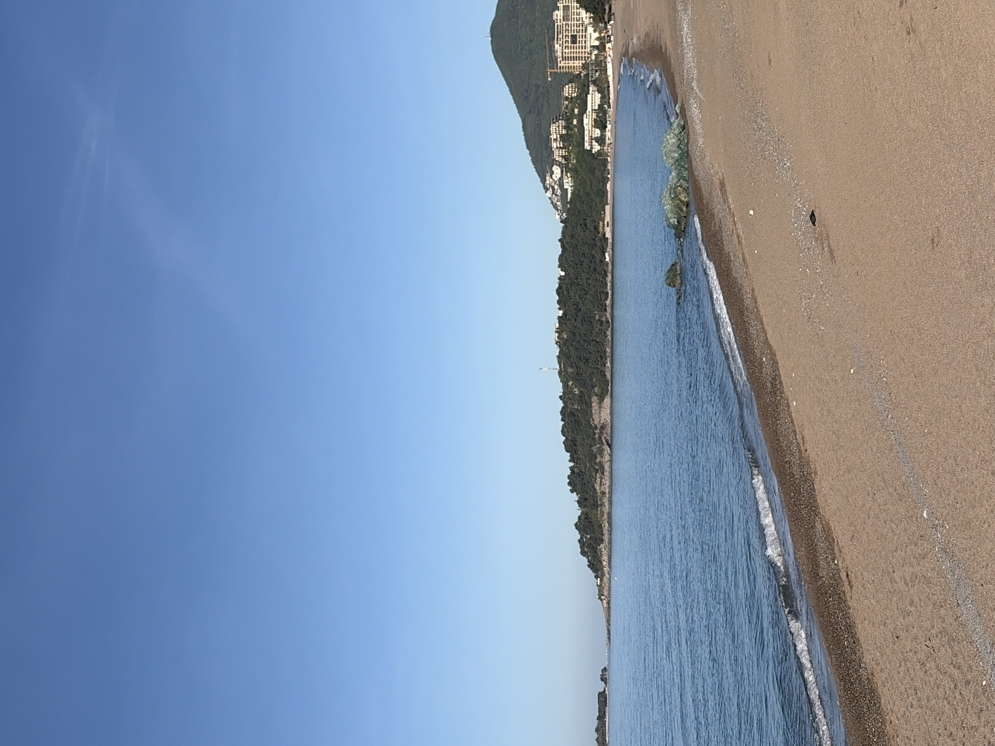 The image depicts a beach in Rafailovići, Montenegro. The scene includes a sandy beach, blue sea, and a clear blue sky. On the right is the sandy beach with a dark brown line of small stones separating it from the sea. The sea has small waves. In the distance, there is a mountain covered in trees and buildings. The sky is cloudless.