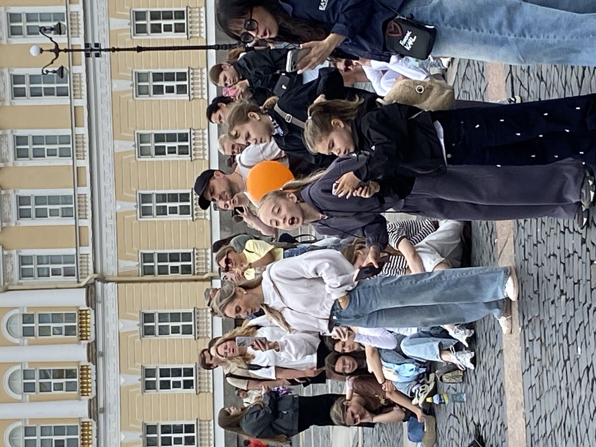 The image depicts a crowd of people gathered in an outdoor urban plaza, likely in Saint Petersburg, Russia, with a neoclassical building in the background. The building is characterized by its yellow facade, numerous windows with white frames, and decorative architectural elements. The ground is paved with cobblestones.
In the foreground, several individuals are visible, some standing and others seated on the ground. A woman in the center foreground, wearing a white hooded jacket and blue jeans, is holding a smartphone and appears to be looking at it or taking a photo. To her left, a young girl with her mouth open is also holding a smartphone. Next to her, another young girl in a black jacket is looking forward.
Other people in the crowd are seen holding up phones, suggesting they might be recording an event or taking pictures. An orange balloon is visible behind some of the people. A lamppost is positioned to the right of the building. The overall scene appears to be a public gathering, possibly a performance or a demonstration.