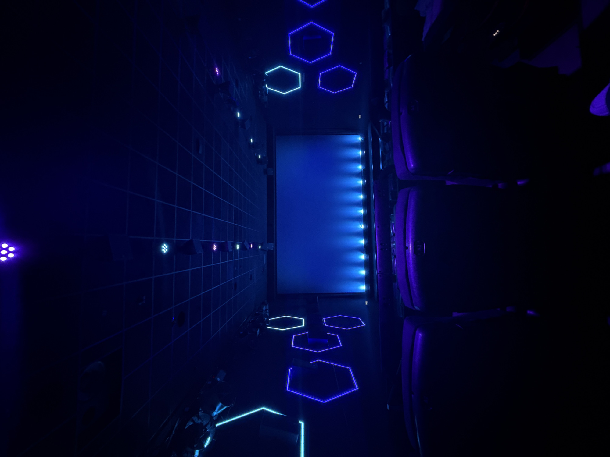 The image depicts an empty cinema hall located in Kassel, Germany, characterized by dark, ambient blue and purple lighting. The scene is observed from a slightly elevated position within the seating area, looking towards the front screen. A large, rectangular projection screen occupies the central front, illuminated from its base by a series of blue lights. The seating area consists of multiple rows of unoccupied, dark-colored (appearing deep purple due to lighting) cinema seats, extending into the foreground and sides. No individuals are present. The side walls of the hall are adorned with various illuminated hexagonal outlines, glowing in shades of blue, purple, and cyan. The ceiling features a dark, gridded structure with recessed lighting fixtures that emit purple, blue, and cyan light. Speakers or sound equipment are subtly integrated into the upper wall sections. The overall ambiance is modern and futuristic, created by the specific color palette and design elements.