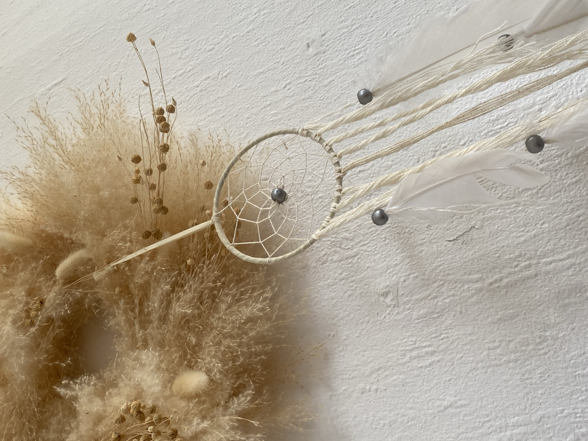 The image shows a dreamcatcher hanging on a textured white wall. The dreamcatcher has a light-colored hoop with a web pattern inside, adorned with several dark grey beads. From the hoop, several strands of off-white string hang down, some with white feathers and more grey beads attached. To the left of the dreamcatcher, a large, fluffy, dried floral arrangement is visible, composed of various dried grasses and seed pods in shades of beige and tan. The arrangement appears to be a bohemian-style wall hanging. The scene is indoors, and the lighting suggests a natural light source from the left, casting subtle shadows. There are no people or animals present. The overall aesthetic is natural, neutral, and bohemian. This scene is not located in the city of Bad Sachsa, Germany, as there is no context or visual information to support that location.