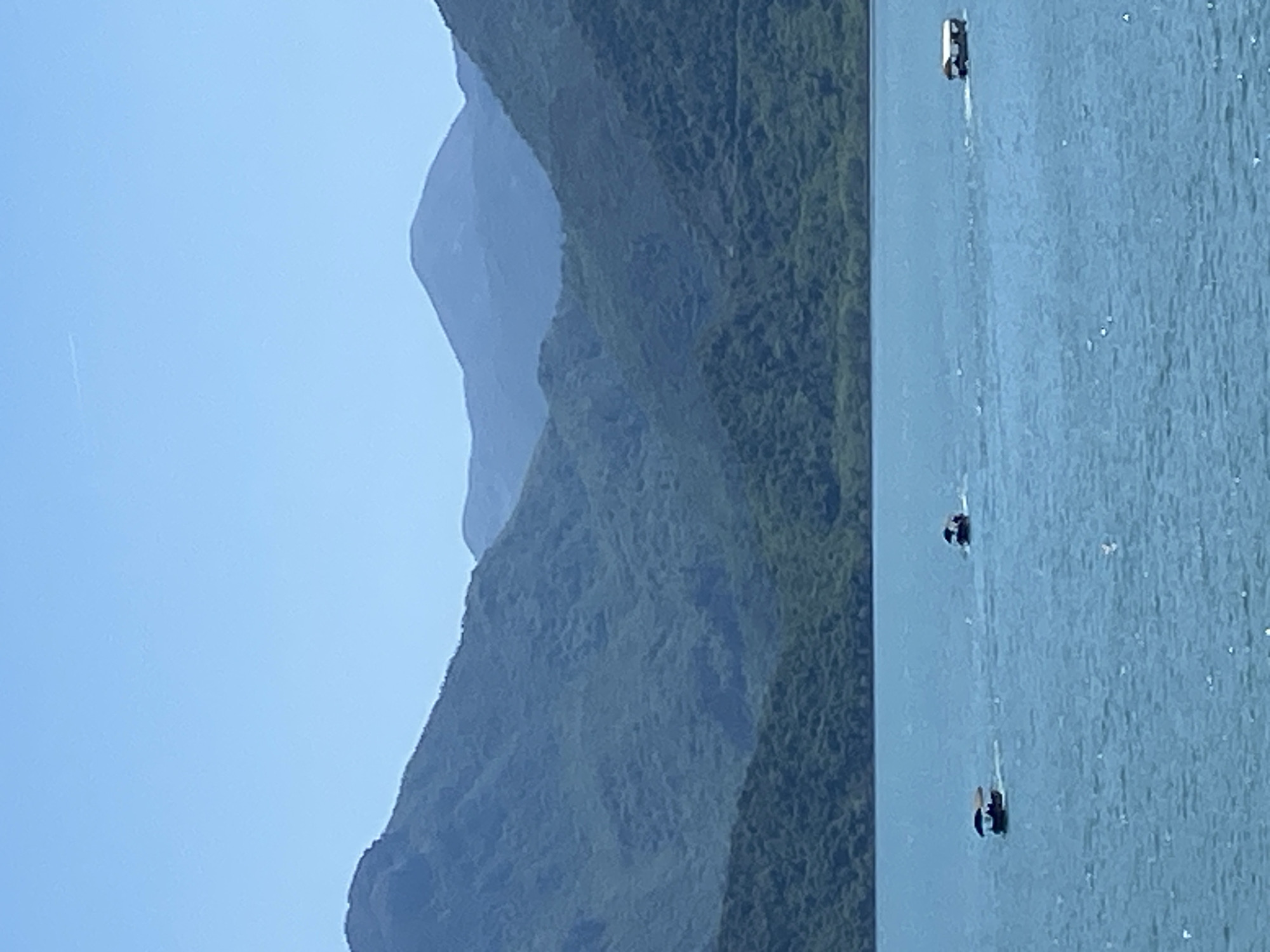 The image shows a landscape with mountains and a body of water, likely a lake or sea. On the right side of the image, boats can be seen on the water. The mountain range in the background features lush vegetation. The sky is clear. The location is plausibly near Kruševica, Montenegro, based on landscape features in that region.