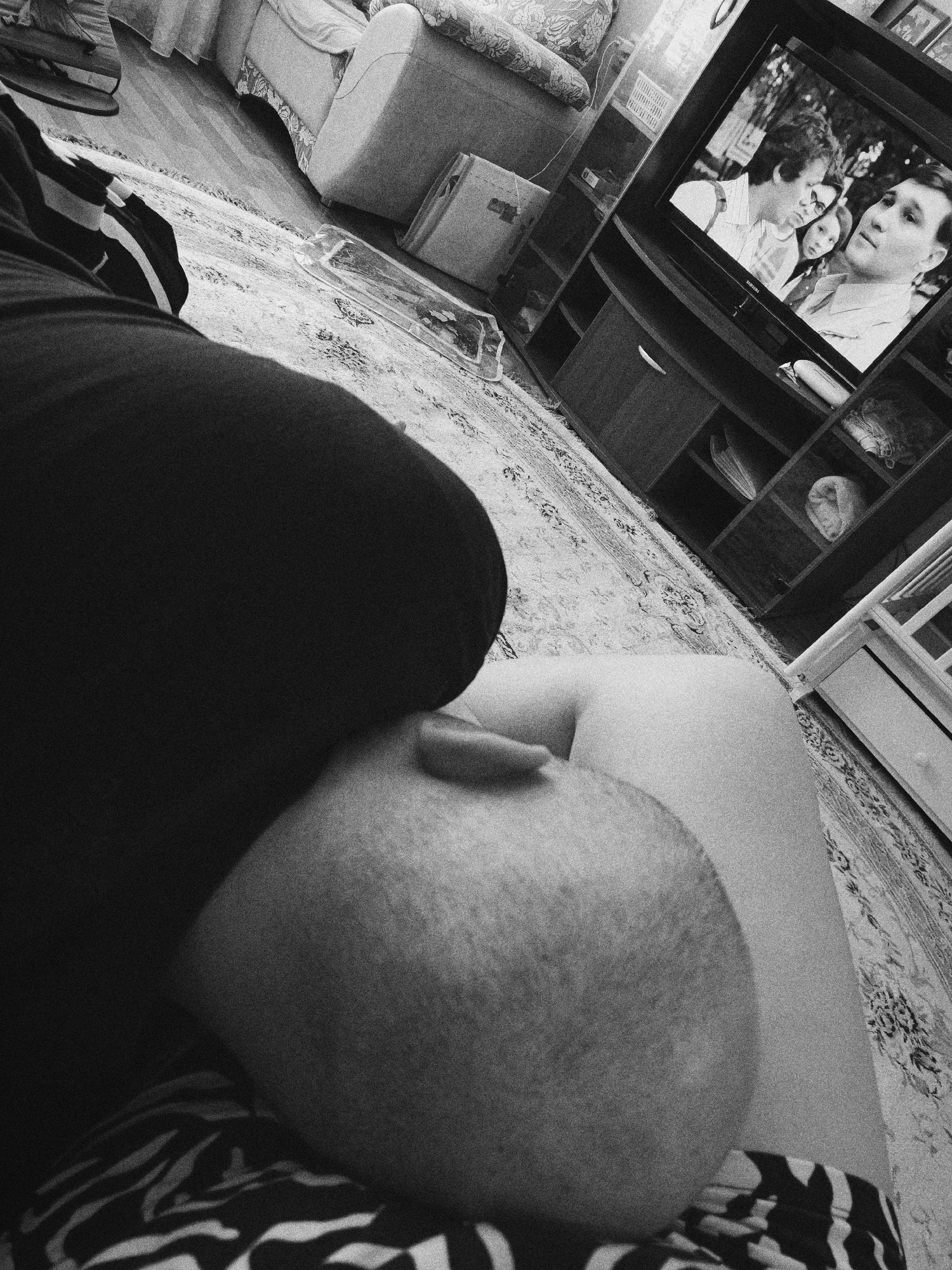 The image shows a person seated in a room. The person's head and upper body are visible. The room contains a sofa, a television displaying a scene with people, and a rug. The exact location within the city of Tobolsk, Russia, cannot be determined from the image.