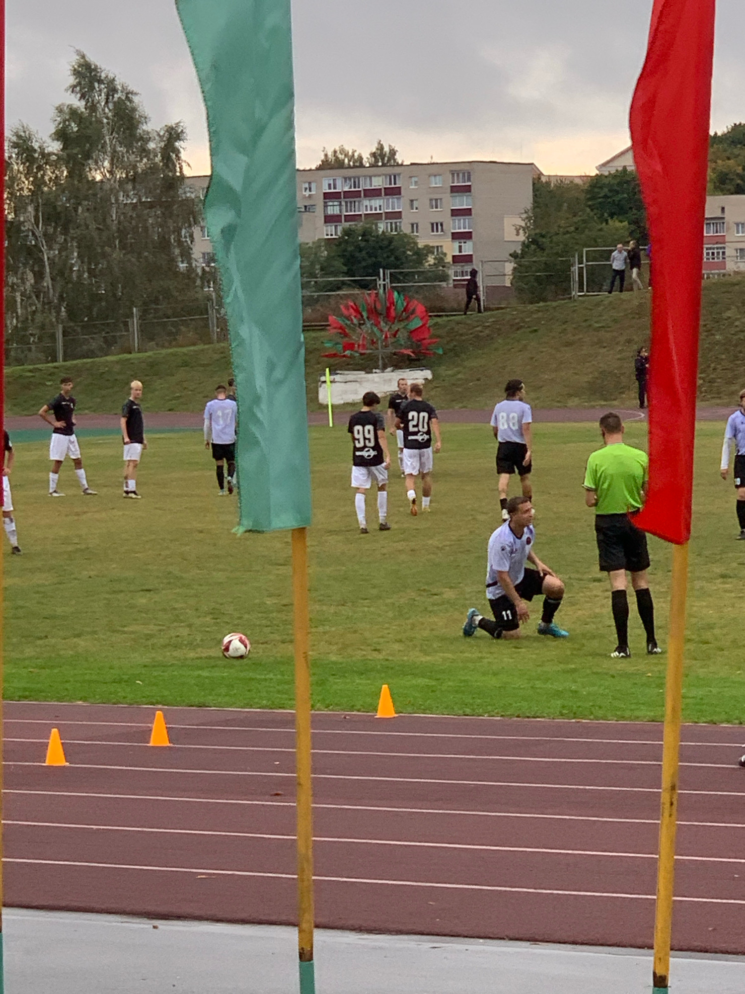 This media file depicts an outdoor soccer match on a grass field, likely a stadium, as indicated by the running track visible in the foreground. The setting appears to be Kapyĺ, Belarus.
The subjects are primarily a group of young men, presumably soccer players, dressed in team uniforms. One player in a light blue uniform is kneeling on the field, wearing the number 11. Another player in a green shirt and black shorts, likely a referee, stands nearby. Other players in black and white, and black and light blue uniforms are scattered across the field, some in action and others observing. A soccer ball is visible on the grass, suggesting an ongoing game or practice.
The event taking place is a soccer match or training session. The flags, one green and one red, suggest a formal sporting event. Orange cones are placed on the running track, possibly for training drills or to mark boundaries. In the background, multi-story apartment buildings are visible, typical of a residential area, and trees line the horizon.
The time of day appears to be late afternoon or early evening, with a cloudy sky casting a diffused light. The weather seems mild, suitable for outdoor sports. There are no overt emotional expressions captured, but the focus and postures of the players suggest a competitive or training environment.
Visible text includes player numbers on uniforms: "11", "20", "88", and "99". The context of Kapyĺ, Belarus, provides a specific geographical location for this sporting scene.