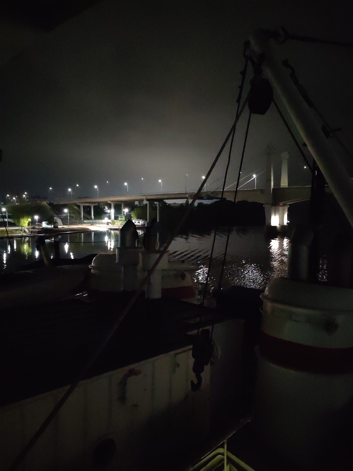 The image depicts a scene at night, seemingly taken from a vessel on a body of water. A bridge is visible in the background, illuminated by streetlights, with its reflection appearing on the water's surface. The foreground is dominated by parts of the vessel, including rigging and what appears to be a white painted structure with a red stripe. The lighting is dim, highlighting the reflections of the bridge lights on the water. Based on the bridge structure, this image appears to be in Kimry, Russia.