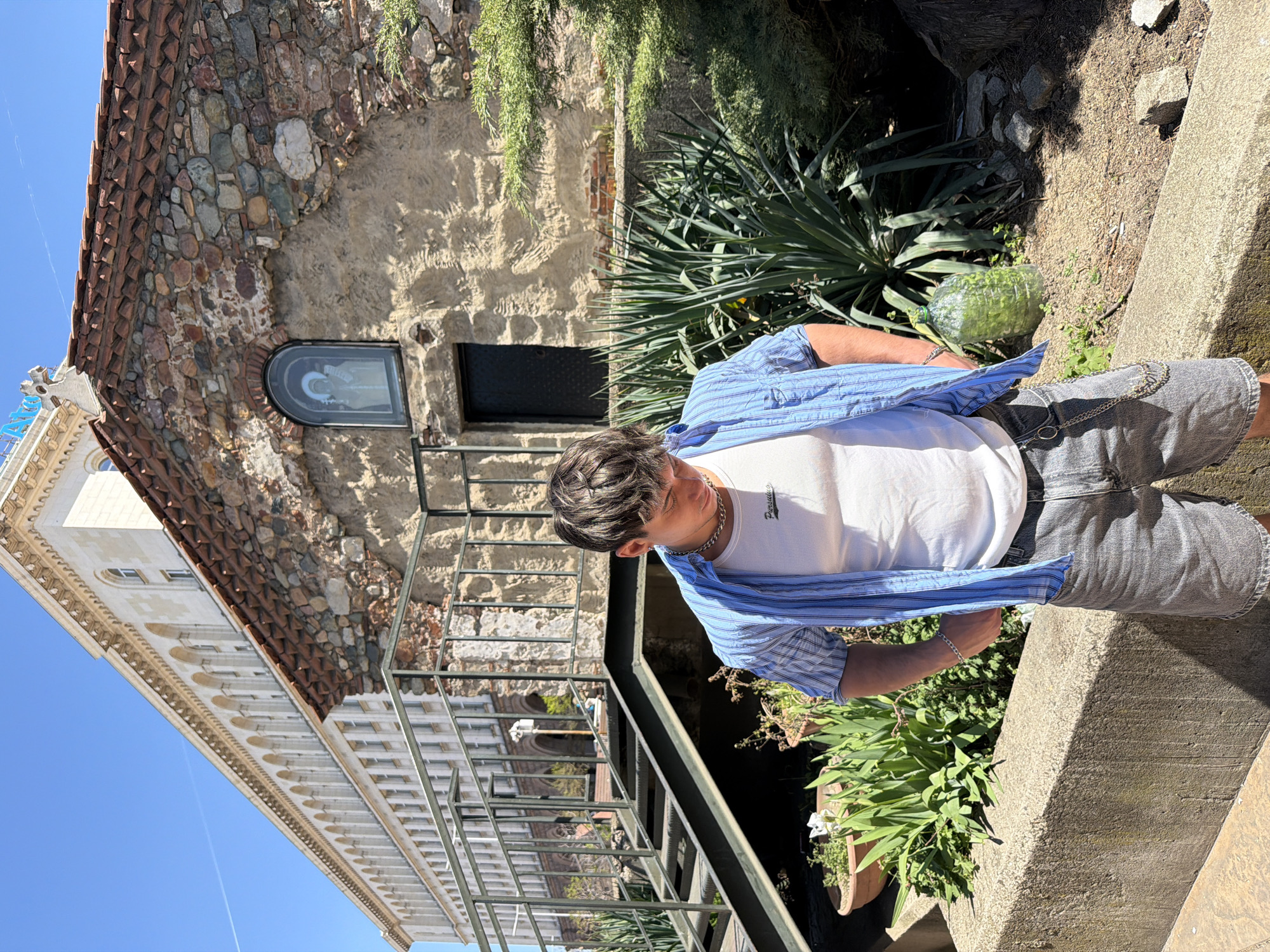 The photograph captures a young man standing beside a concrete planter, set against the backdrop of a historic building in Sofia, Bulgaria. The man is dressed in a white t-shirt, over which he wears a blue striped shirt. Gray shorts and a chain accessory complete his casual outfit. Behind him, a multi-story building is visible, constructed with a combination of stone and tile. The architectural design includes arched windows and decorative trim. A metal staircase is positioned adjacent to the building, leading to an upper level. The sky overhead is clear and blue. The scene suggests an outdoor setting, potentially a public space or garden near the architectural structure. The light is bright and creates strong shadows.