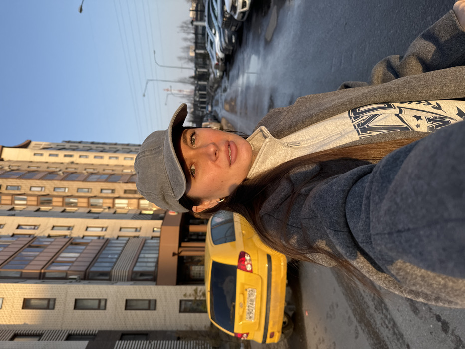 A female subject, estimated to be in her 20s or 30s, is depicted in a selfie, extending her left arm toward the camera. She wears a grey baseball cap, a light grey quarter-zip top, and a dark grey long coat. Her long, dark hair is visible beneath the cap, and she has small silver hoop earrings. The scene is set on a wet, paved street or parking area in the city of Odintsovo, Russia.
Behind the subject, to her left, a yellow Hyundai Getz is parked, displaying Russian license plate H907AB 159. Immediately behind the vehicle, a modern multi-story residential building with a light-toned brick or stone facade and dark windows rises. The building's surfaces are illuminated by low-angle sunlight.
To the subject's right, the wet pavement extends into the background, flanked by a row of numerous parked passenger vehicles. Further in the distance, streetlights are visible. Beyond the street and cars, a fence separates the area from industrial or utility structures, including a tall, slender column resembling a chimney or power line tower, silhouetted against a clear blue sky. The ground across the visible area is reflective due to moisture, suggesting recent precipitation. The overall lighting conditions indicate either early morning or late afternoon.