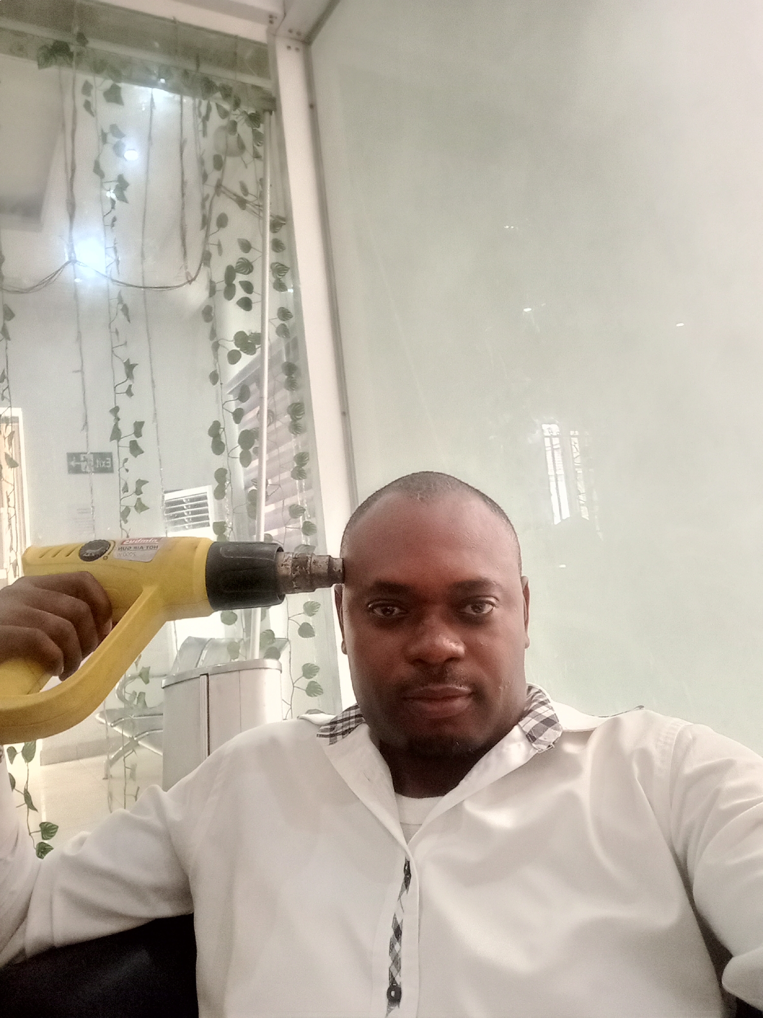 The image shows a man in Gwarinpa, Nigeria holding a yellow heat gun to his head, possibly in jest. He is wearing a white button-down shirt with a patterned collar. The background includes a window with decorative ivy and a white surface, potentially a wall or panel. There is also a white filing cabinet behind him.
