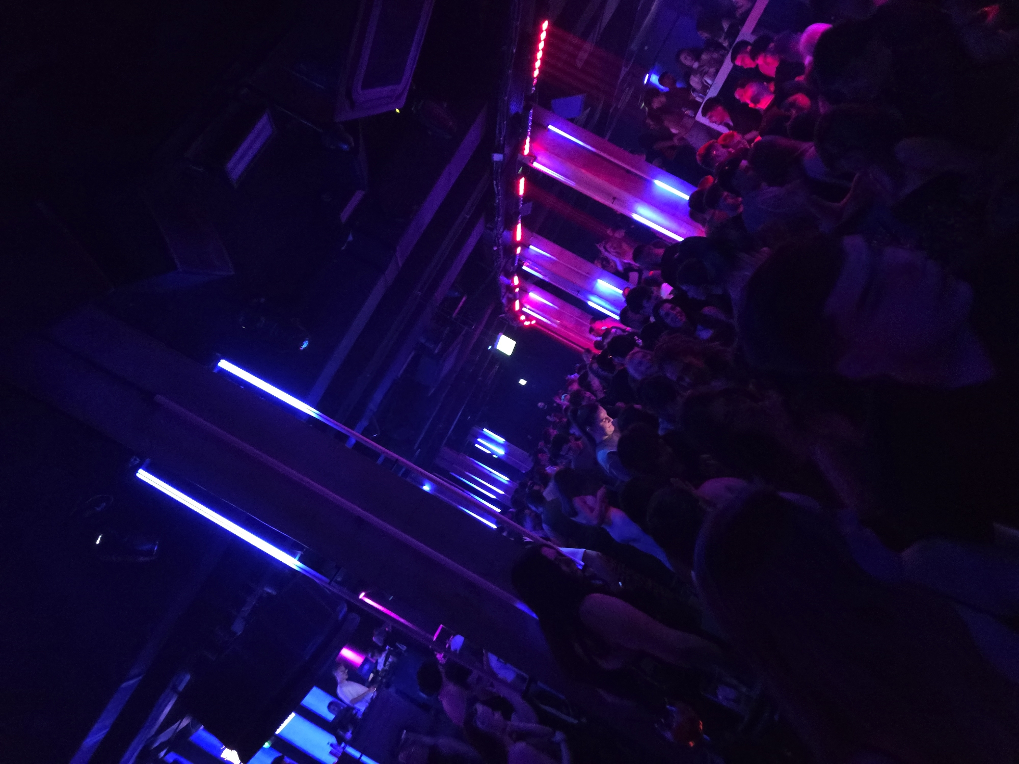 The image captures a nightclub scene, possibly in Varna, Bulgaria, as suggested by the prompt. A dense crowd of people is present, illuminated by purple and red neon lights. The location appears to be a dance floor or concert venue based on the arrangement of lighting. The photograph is tilted, making it difficult to discern the precise positioning of the subjects, but the atmosphere is suggestive of a crowded, high-energy nightlife setting.
