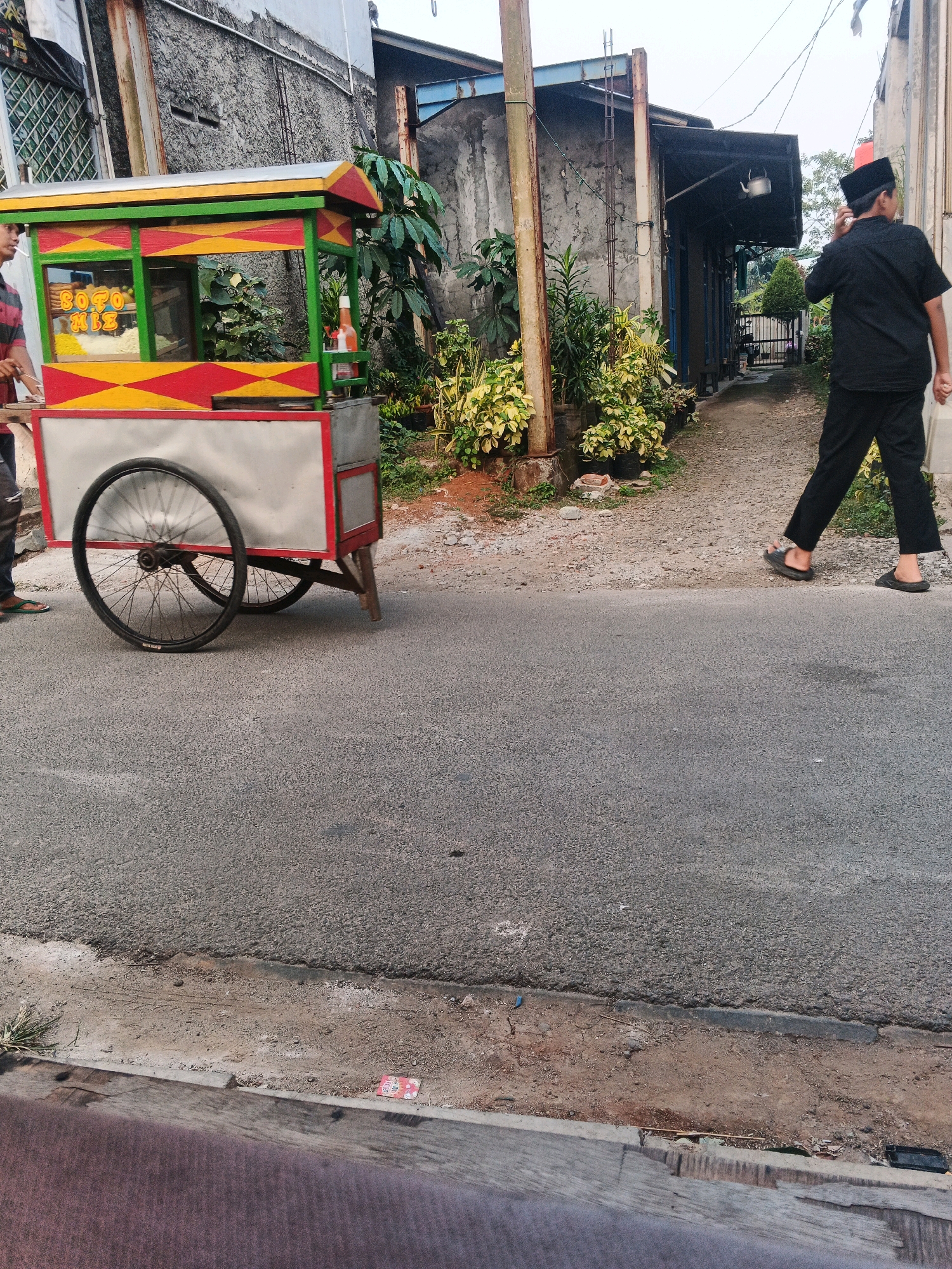 In Kadu, Indonesia, a street scene unfolds with a colorful food cart dominating the left side of the frame. The cart, painted in vibrant red, yellow, and green, displays the text "SOTO MBZ" on its side, suggesting it sells Soto, a traditional Indonesian soup. Inside the cart, various food items are visible, and a bottle of red sauce sits on the right. A person in a striped shirt stands by the cart, partially obscured, with their left side visible.
To the right, a man dressed in a black shirt and pants, and wearing a black peci (a cap worn by Muslim men in Southeast Asia), walks away from the cart and towards a building in the background. He is seen with his hand near his face, as if adjusting his cap or looking at something. The background features buildings with textured walls, some with metal roofing. Lush green plants and potted plants are scattered along the pathway leading to the buildings. The ground is a mix of asphalt and dirt. The overall scene captures a slice of daily life on a street in Kadu.