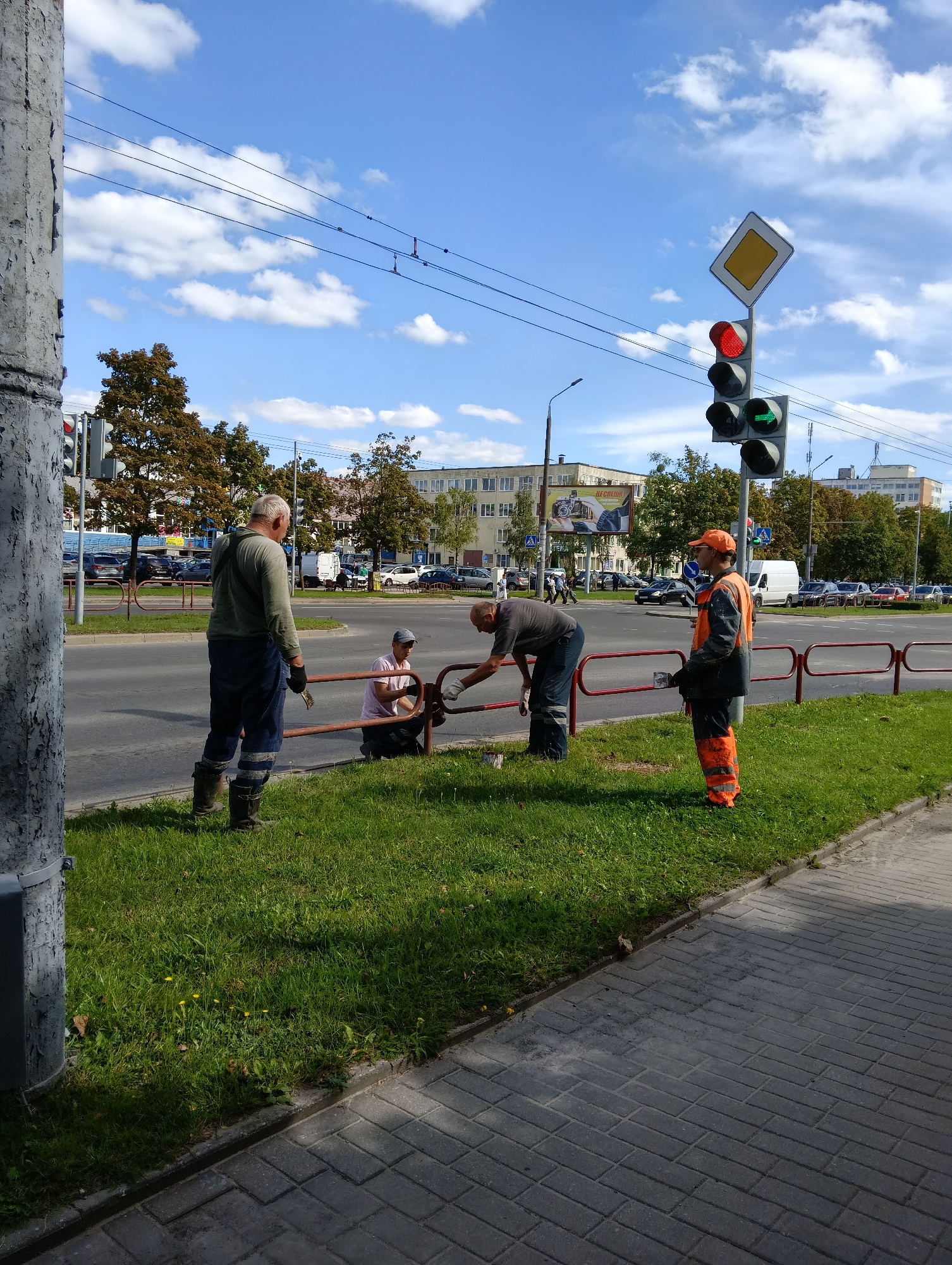 In Hrodna, Belarus, three individuals are engaged in work near a roadside on a sunny day. Two men, wearing work attire including overalls and boots, are near a red metal railing. One man, with gray hair, stands and holds a cleaning rag, while another man, wearing a cap, is kneeling and appears to be cleaning or repairing the railing. A third person, dressed in a high-visibility orange uniform, stands facing the railing, holding a tool or object in their right hand. They are positioned on a grassy area adjacent to a paved sidewalk and a street with parked cars and a traffic light visible in the background. The traffic light shows a red signal in the top position and a green arrow signal in the bottom position. Power lines and a diamond-shaped road sign are also visible above the traffic light. The sky is blue with scattered white clouds.