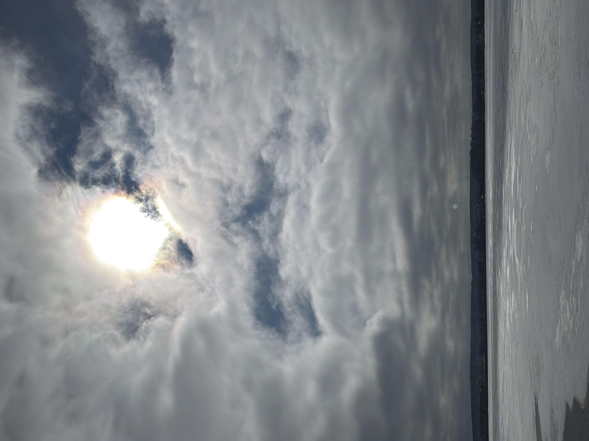 The image captures a cloudy sky over what appears to be a frozen body of water. The sun is obscured by the clouds, creating a bright spot with a slight rainbow effect. A line of trees is visible along the horizon where the sky meets the water. The water appears to be frozen with a smooth, slightly reflective surface. Given the context provided, this scene is likely a view over the Votkinsk Reservoir in Votkinsk, Russia, during a period of cold weather when the reservoir is frozen. No people are discernible in the image.