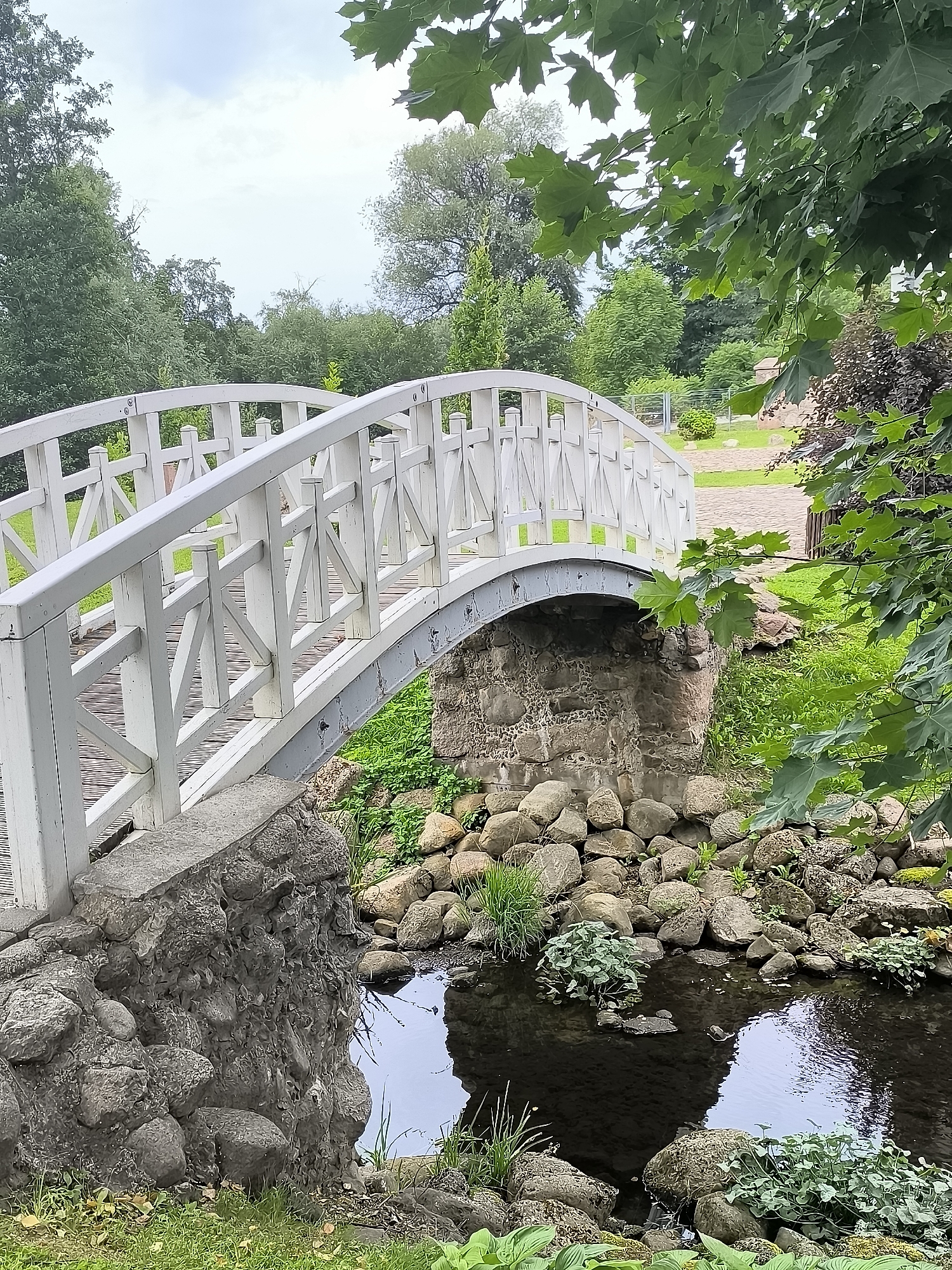 The image shows a white wooden pedestrian bridge arching over a small stream. The bridge has a lattice railing and a metal support structure underneath. The stream banks are lined with large rocks and vegetation. In the background, there are trees and a paved area. The scene is set in a park-like environment, likely in Kretinga, Lithuania, given the prompt's context. There are no people visible in the image. The sky is overcast.